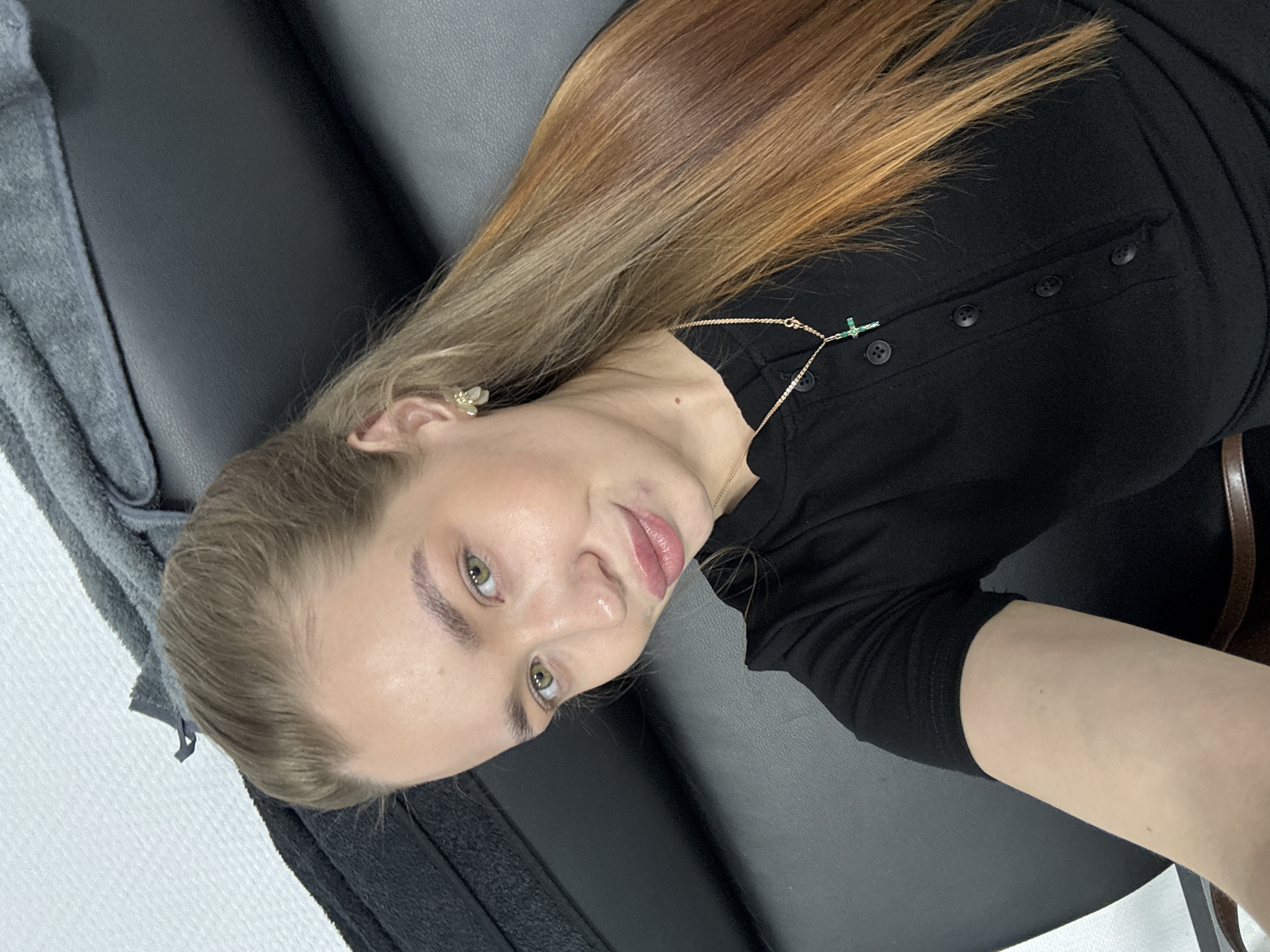 A woman with long, light brown to reddish-brown hair is lying on a black, smooth surface, possibly a padded table or bench. She is wearing a black button-down shirt and a delicate gold necklace with a small green cross pendant. She has fair skin, light eyes, and is looking directly at the camera with a slight smile. To the left of her head is a gray, textured surface, possibly a towel or blanket. The background is a plain white wall. The location is Chaikovsky, Russia.
