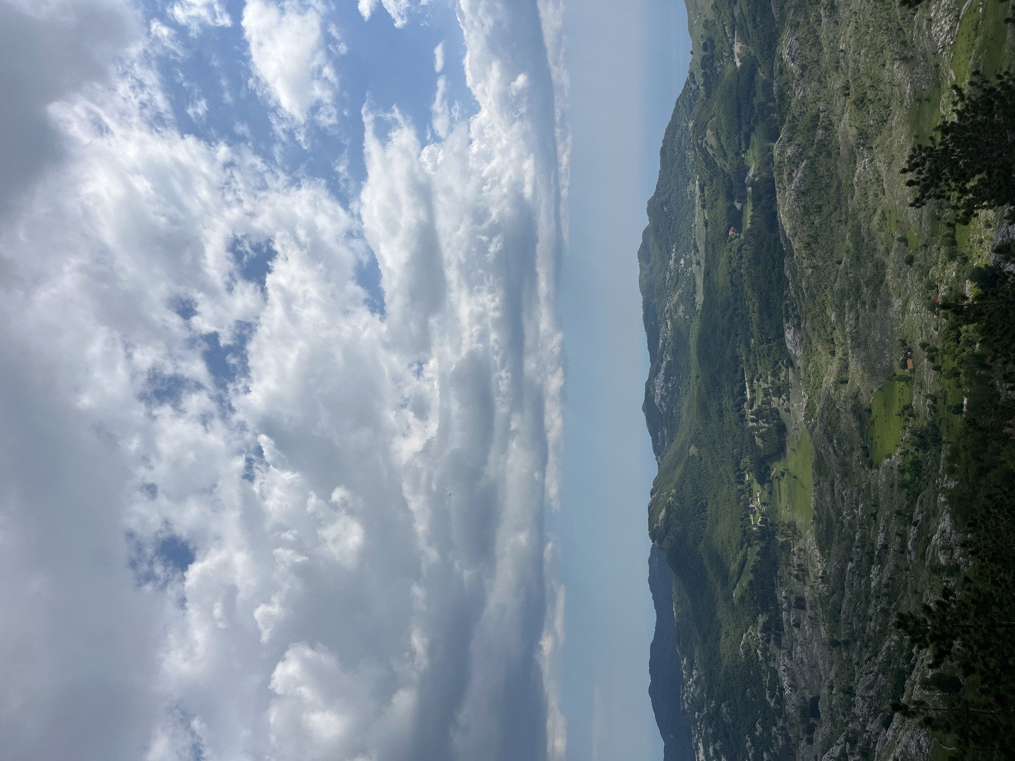 The image shows a landscape in Dolovi, Montenegro. On the right, a mountain range covered with green vegetation and rocky terrain is visible. A few buildings and fields are scattered on the mountainside. The sky on the left is filled with various cloud formations, ranging from fluffy white clouds to darker, more dense patches. In between the mountain and the sky, a band of hazy blue suggests the presence of a horizon or distant atmosphere.