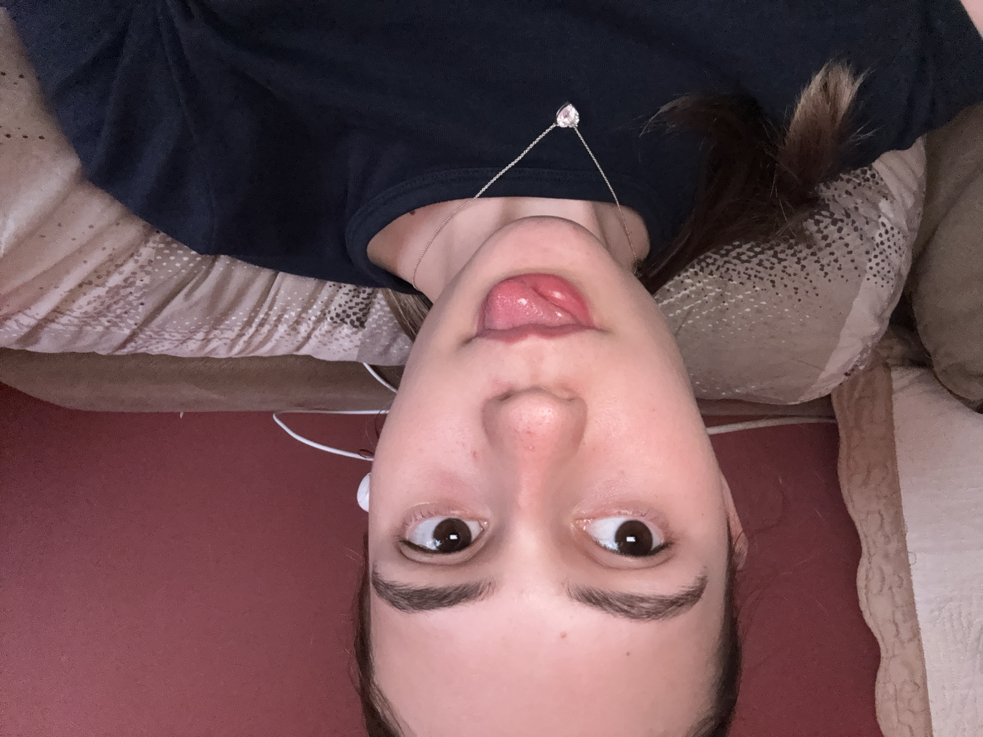 A person with brown hair and brown eyes lies on a bed in a room with red flooring. The person wears a dark shirt and a silver necklace with a heart-shaped pendant. They stick out their tongue and appear to be wearing earphones. The location is not discernible from the image.