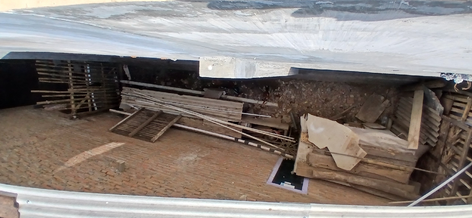 In the city of Pejaten, Indonesia, an exterior view captures a construction site with a brick wall on the left and a light-colored, possibly concrete wall on the right. The area between these walls appears to be a narrow alley or courtyard. The ground is covered with debris, including dirt, rocks, and various construction materials such as wooden planks, pipes, and corrugated metal sheets. A wooden grate or frame is visible on the ground towards the bottom of the frame. In the background, a makeshift fence made of vertical wooden stakes and horizontal supports is erected. A small, dark rectangular opening, possibly a window, is visible on the brick wall. The scene is tilted, suggesting the photograph was taken from an elevated perspective.