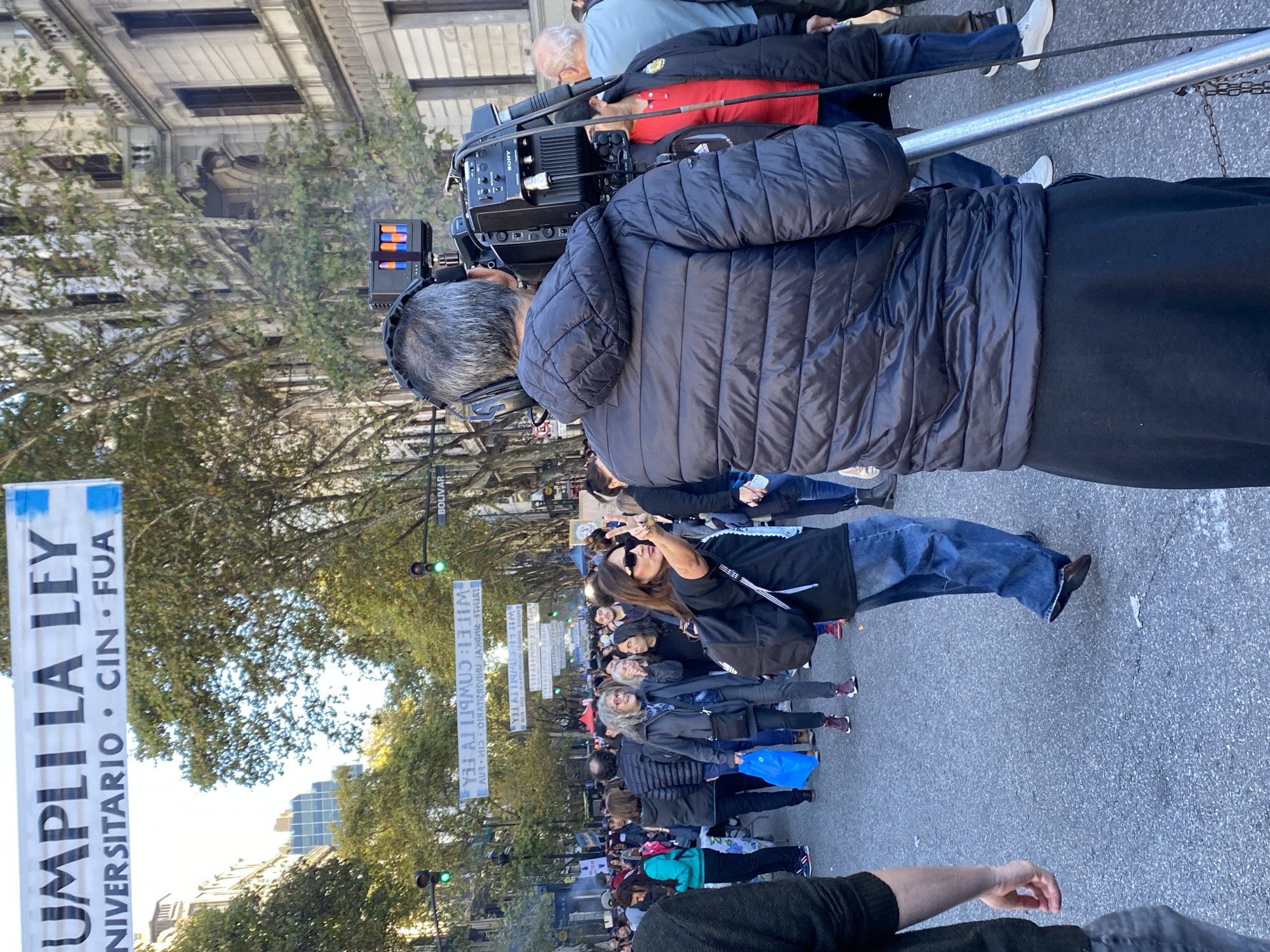 The image depicts a rally or demonstration in Buenos Aires, Argentina. A crowd of people is moving along a street named Bolivar, holding banners that read "UMPLI LA LEY UNIVERSITARIO CIN FUA" (Comply with the University Law, CIN FUA). A camera crew films the event, while people in the crowd are taking photographs. Buildings can be seen in the background, along with trees. The traffic lights on Bolivar Street are green. The overall atmosphere appears to be one of protest or advocacy.