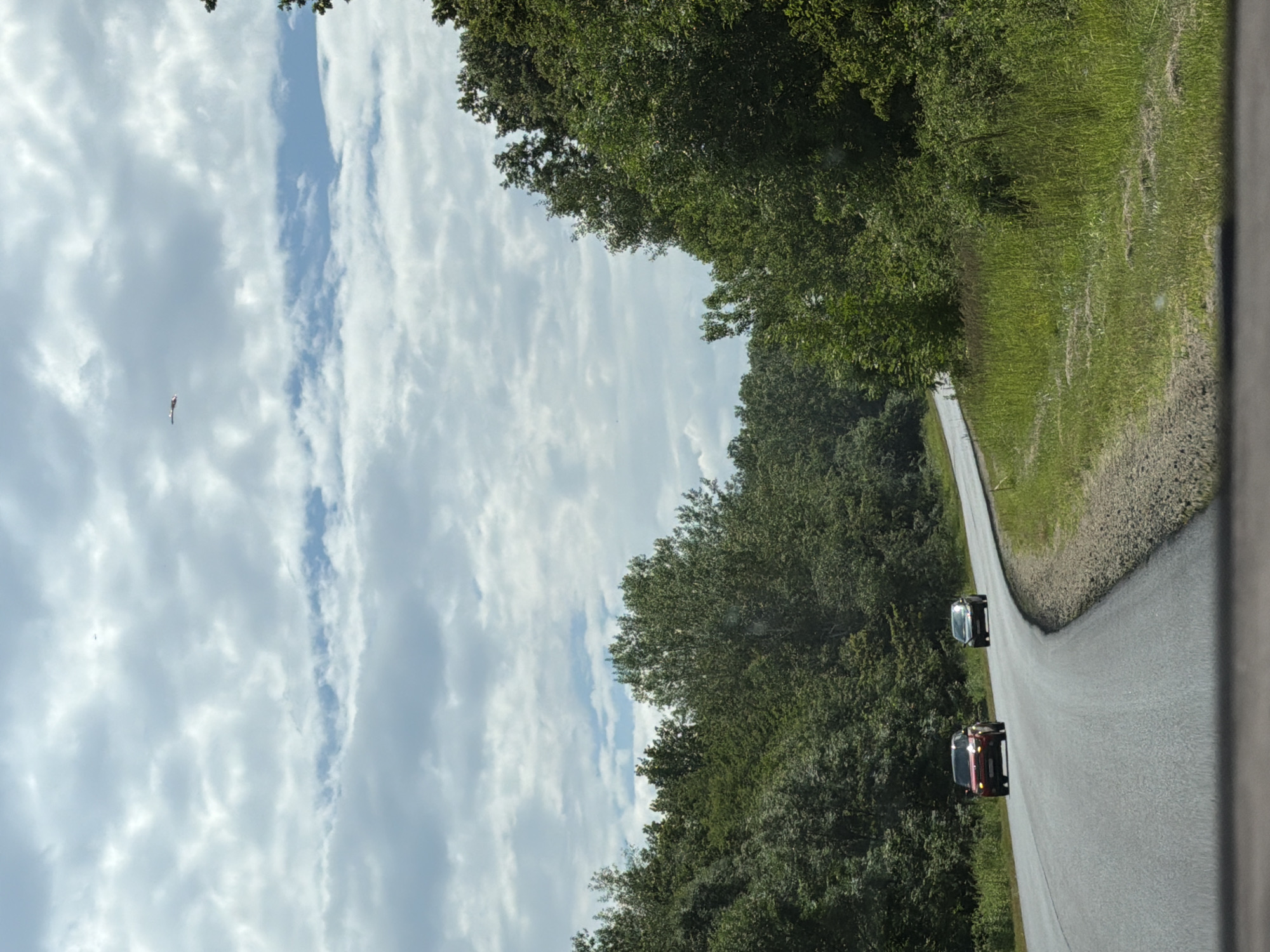 The image shows a highway with two cars driving on it, surrounded by lush green trees and a cloudy sky. In the sky, a small, distant airplane is visible. The scene appears to be a rural or suburban area, possibly on a road leading to or from Krasnooktiabrskiy, Russia, given the typical landscape and infrastructure of the region. The weather is overcast, suggesting a partly cloudy day. The cars are positioned on the road in a way that suggests normal traffic flow. The trees on either side of the road indicate a natural, wooded environment.