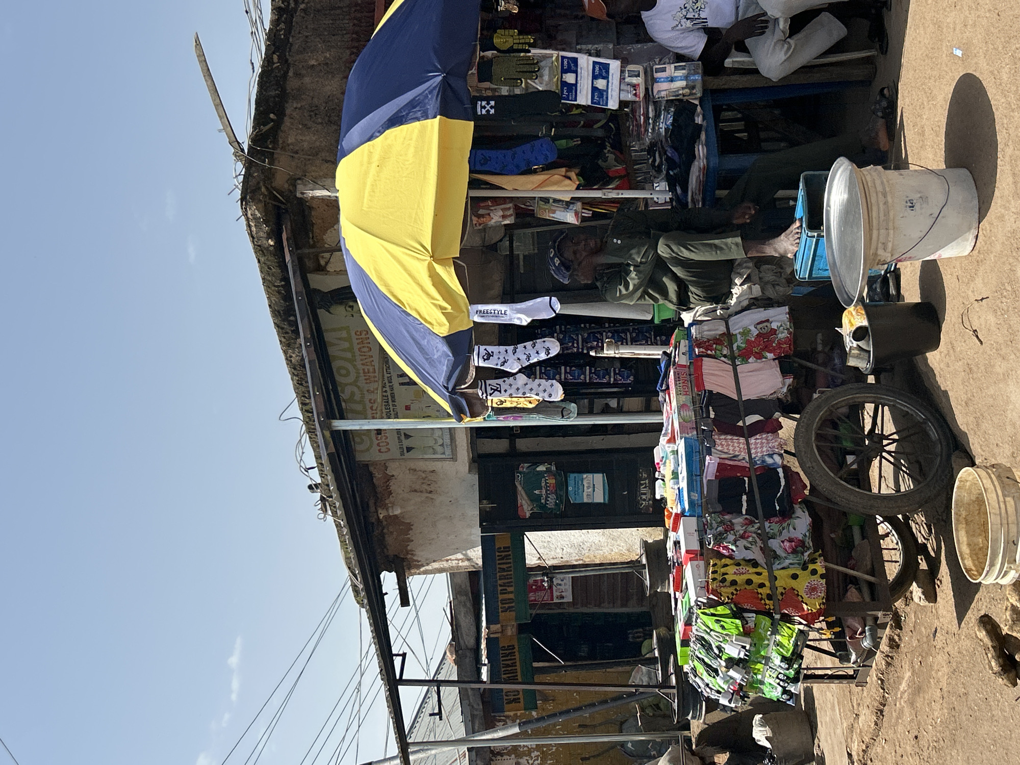 This media file shows an outdoor market stall on a sunny day. A vendor is seated behind a display of clothing and accessories, including socks and what appear to be wigs. The stall is adorned with a large blue and yellow umbrella for shade. In the foreground, a cart is piled high with colorful items. A white bucket and a black bin are also visible. The scene appears to be located in a dusty, urban environment, with a building in the background. Signs on the building and stall provide some textual information, including "NO PARKING" and "COSMISS WEAVONS". The overall impression is one of a bustling marketplace.