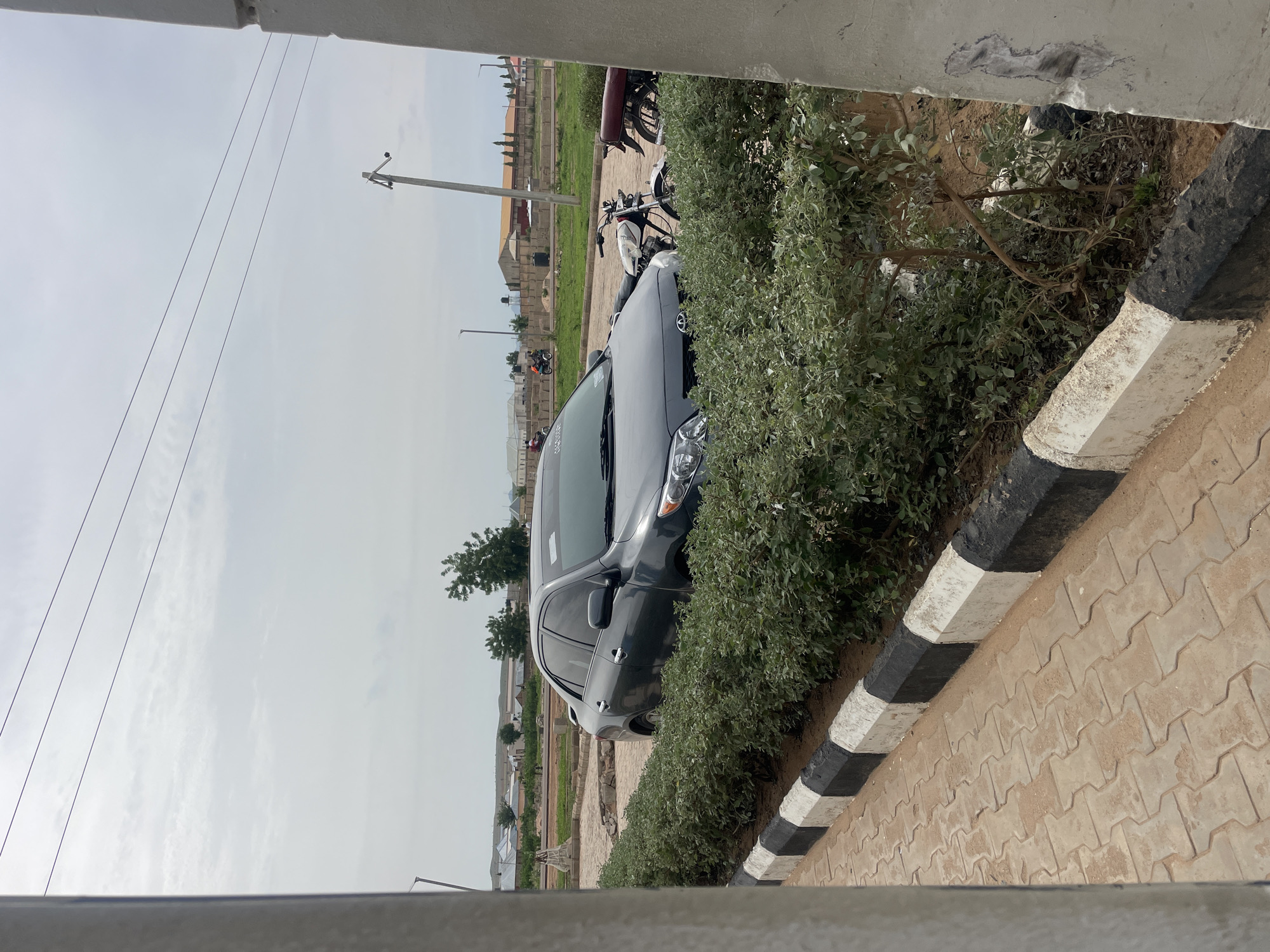 This daytime image, set in Pindiga, Nigeria, captures an outdoor scene under an overcast or hazy sky. The central subject is a dark grey sedan, possibly a Toyota, parked next to a vibrant green, well-trimmed hedge. The car's front passenger side, including its headlight and windshield, is clearly visible.
The hedge runs parallel to a paved area constructed from light-colored, interlocking concrete pavers. A distinct curb, painted with alternating black and white stripes, separates the hedge from the pathway. In the background, behind the hedge and the parked car, several motorcycles are visible, including what appears to be a dirt bike or scooter.
The far background reveals an expanse of undeveloped land dotted with scattered, low-rise buildings and what might be ongoing construction or foundations, indicative of a developing or semi-urban environment. Utility poles and multiple power lines stretch across the sky. The photograph seems to be taken from a slightly elevated vantage point, possibly from a building or structure, as suggested by the concrete framing elements visible at the top and bottom edges of the image. No people or legible text are present in the scene, which conveys a sense of stillness and calm.