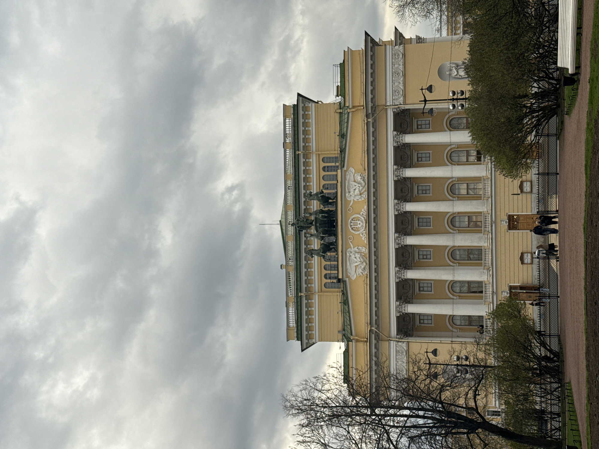 The image depicts the Alexandrinsky Theatre in Saint Petersburg, Russia, viewed from a slightly low angle. The building is a classical structure with a yellow facade, white columns, and ornate sculptures. A quadriga statue adorns the top of the building. To the right of the building is a park or garden area with trees and a path. Several people can be seen walking along the path. The sky is overcast with grey clouds.