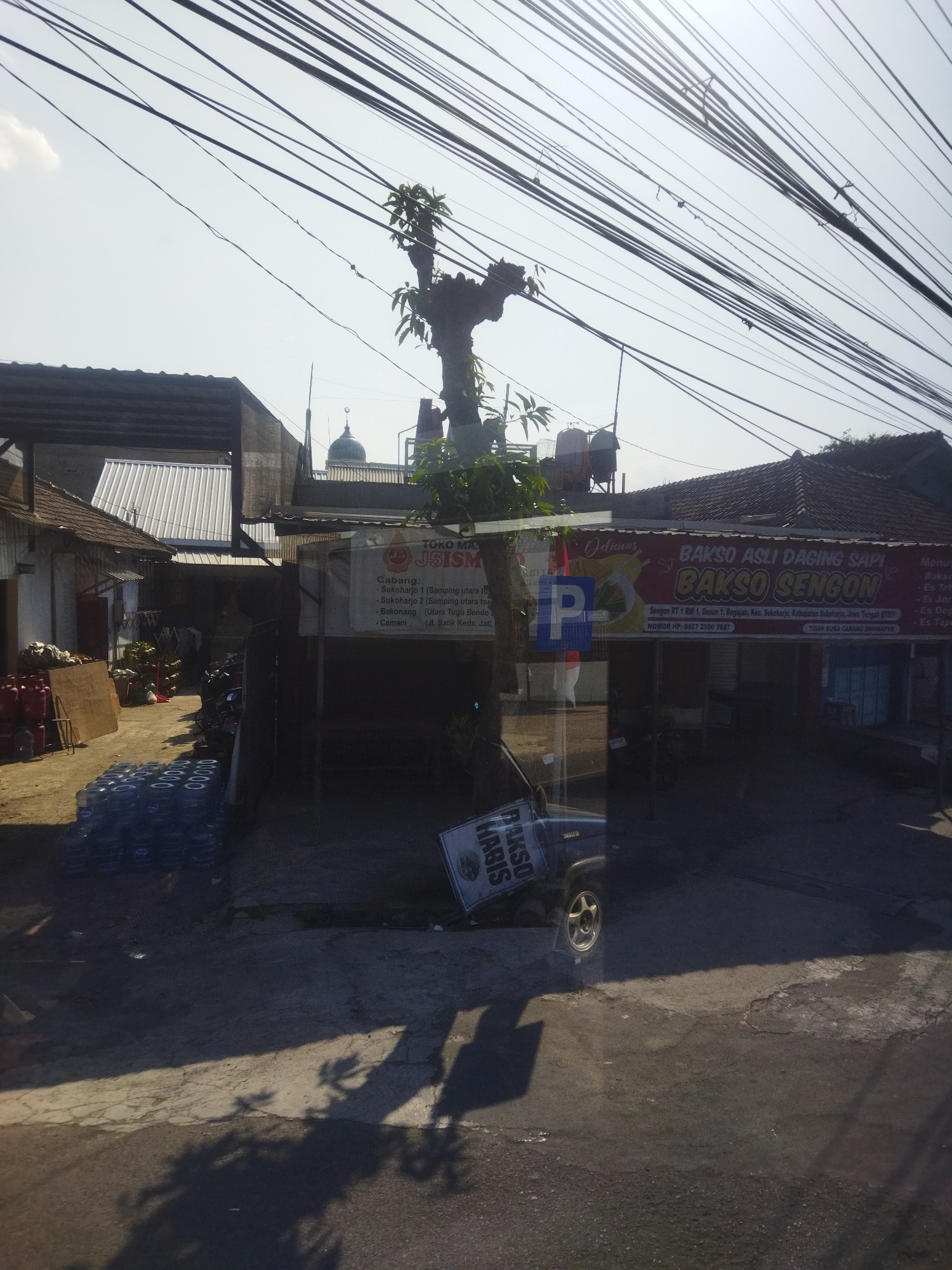 The image depicts a street scene in the city of Begajah, Indonesia, with a focus on a building with a red awning and signage. Overhead, a dense network of electrical wires crisscrosses the sky. The building's awning reads "BAKSO ASLI DAGING SAPI BAKSO SENGON" and "SEBOKON RT 1 RW 1, Dusun 1, Begajah, Kec. Sukoharjo, Jawa Tengah 57397". Beneath the awning, the interior of the establishment is visible, showing tables and chairs, suggesting it is a food stall or restaurant. To the left of this building, another structure with a corrugated metal roof is present. In front of these buildings, stacked blue water jugs are arranged on the ground, and a dark-colored SUV is parked. The ground is paved with a mix of asphalt and concrete, with shadows cast by overhead trees and structures. The overall impression is of a typical Indonesian street scene.