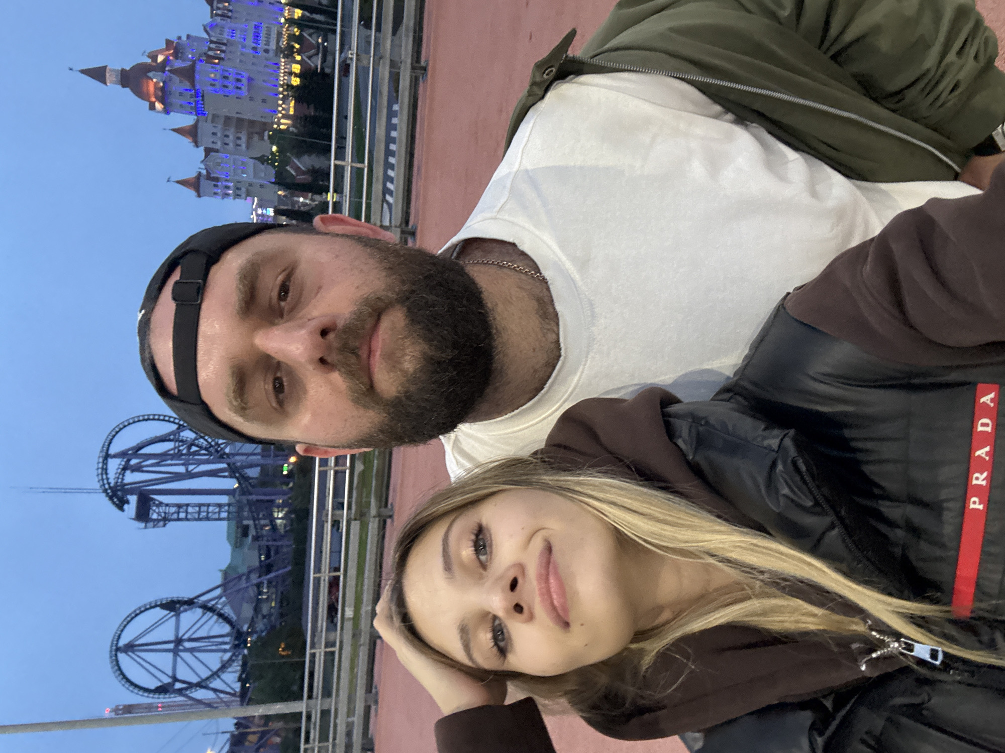The image shows a selfie of a man and a woman, likely taken in Sirius, Russia, given the presence of Sochi Park. The man is fair-skinned, with a beard and a black baseball cap. He is wearing a white t-shirt and an olive green jacket. The woman has light-colored hair and is wearing a brown hooded sweatshirt and a black Prada jacket with a red stripe. In the background is a brightly lit castle-like structure, which may be Sochi Park's Bogatyr Castle Hotel. A rollercoaster structure is also visible in the background, indicative of an amusement park setting. The image appears to have been taken at dusk.