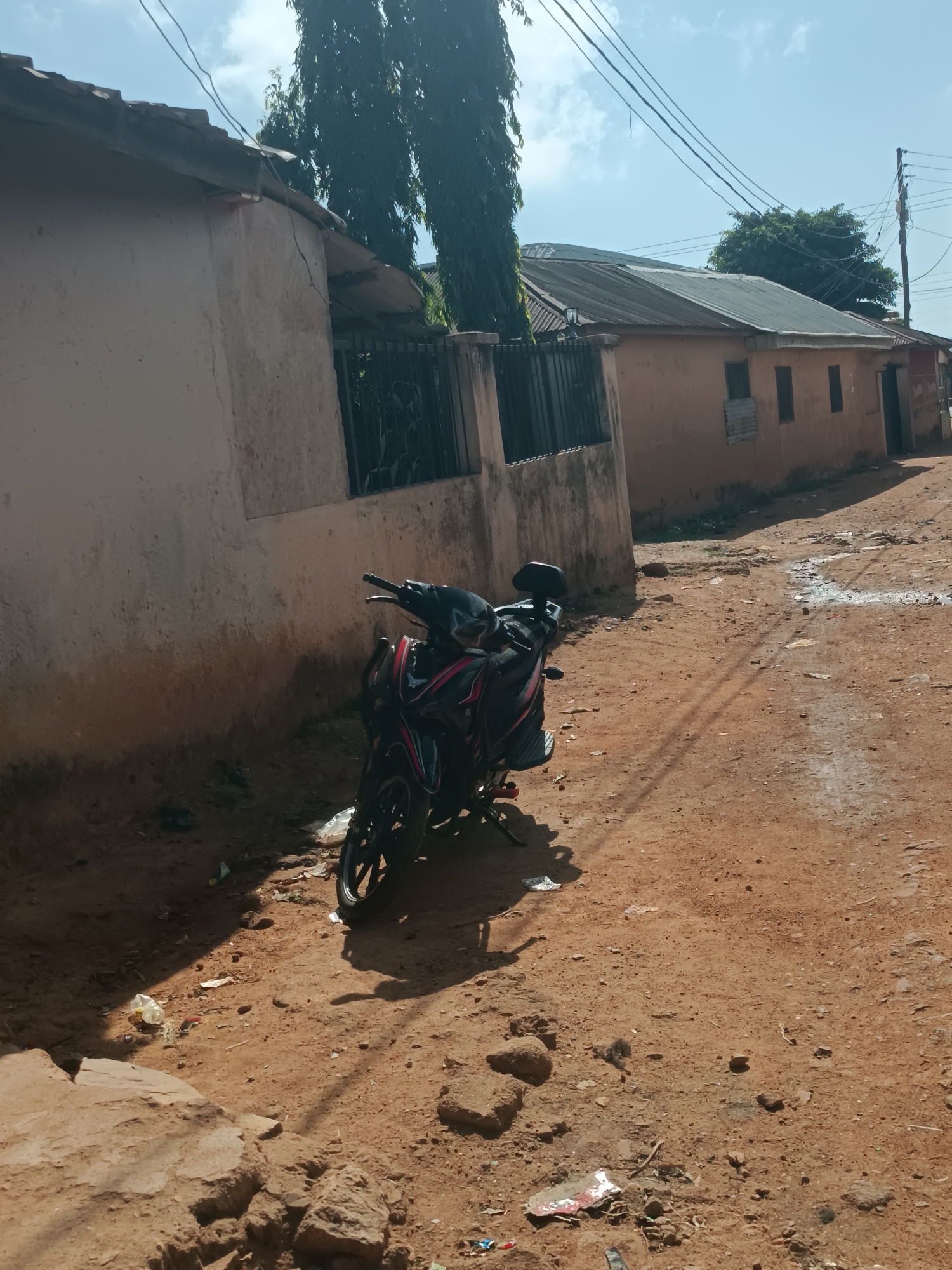 The image shows a motorcycle parked on a dirt road in Bukuru, Nigeria. The road is lined with buildings, one with a fence. Debris litters the ground. The sky is partly cloudy. There are no people visible. Powerlines are strung overhead.