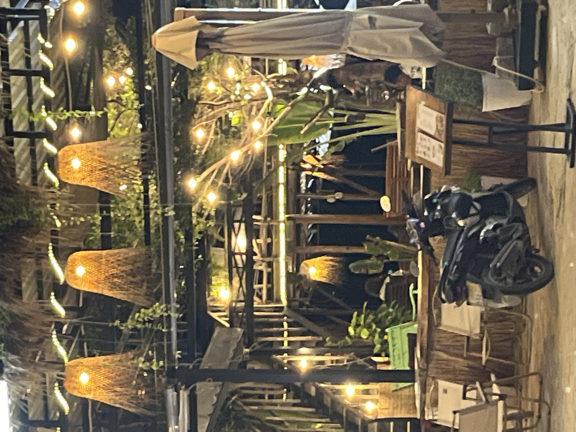 This is a nighttime outdoor scene, possibly a restaurant or cafe, with a rustic, bohemian aesthetic. The setting is filled with warm, ambient lighting from strings of Edison-style bulbs and woven lamp shades that hang like decorative lanterns. Tropical foliage, including large leafy plants, is visible throughout the space, suggesting a warm climate. Wooden structures, possibly part of the cafe's architecture or decor, create a natural and textured environment.
In the foreground, a dark-colored motorcycle is parked, adding a modern element to the otherwise organic and rustic surroundings. Several chairs, some appearing to be made of woven material and others more modern, are arranged around tables, though no patrons are clearly visible in the immediate area. A sign on a wooden easel stands near the motorcycle, displaying what appears to be a menu or information, though the text is illegible.
The overall impression is one of a cozy and inviting evening spot, likely in a tropical or subtropical region. The time of day is clearly night, indicated by the darkness and the prominent artificial lighting. The weather appears calm, as there are no signs of rain or wind. The location context of Phu Thuy Ward, Vietnam, further supports the idea of a warm, possibly humid, evening setting. The scene evokes a relaxed and perhaps romantic atmosphere.