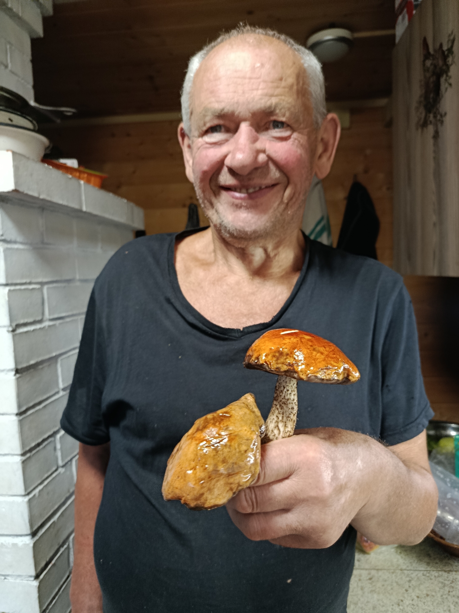 A Caucasian man with short, gray hair and a slight stubble is smiling and holding two mushrooms in his right hand. He is wearing a black t-shirt. The mushrooms have orange caps and textured stems. The background appears to be a rustic interior with wooden walls and a white brick structure. The lighting is dim. The man is positioned in the center of the frame, and the mushrooms are held in the foreground, slightly to the right.