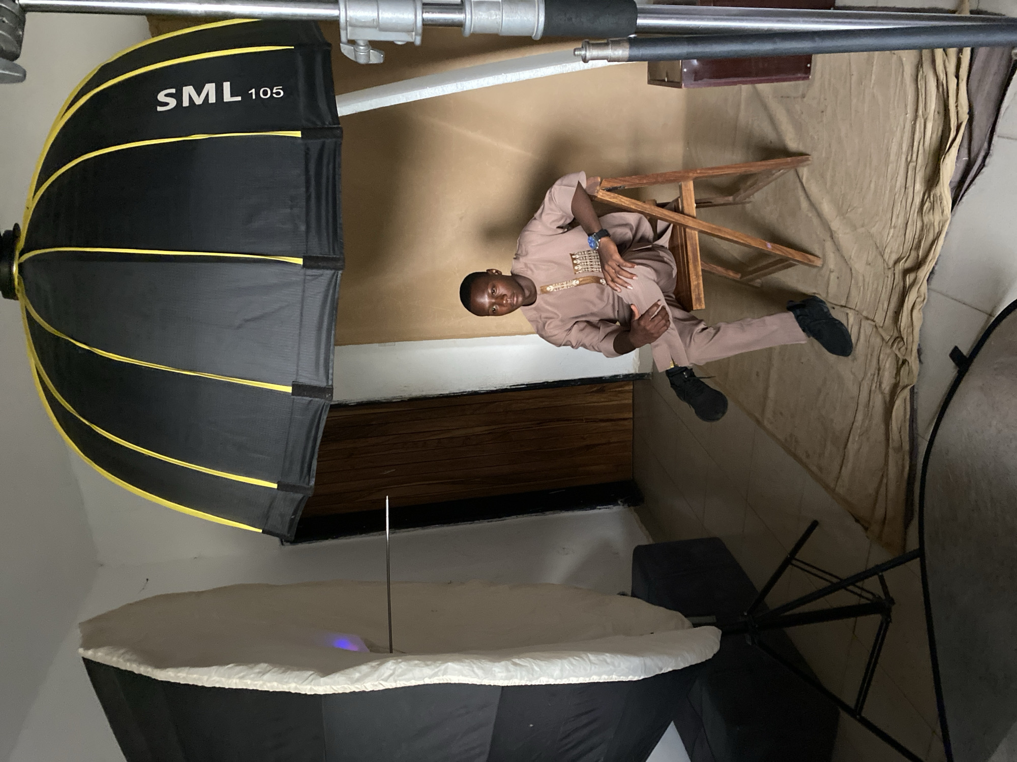 A medium shot depicts a man seated on a wooden chair in what appears to be a photography studio. The man, dressed in a brown traditional attire, sits with his legs crossed and his hands resting on his lap. To the left are two softboxes, one black with yellow trim labeled "SML 105" and the other white. Overhead, studio lighting equipment is visible. The background consists of a neutral backdrop, likely used for portraits or studio photography. The location is Bukuru, Nigeria.