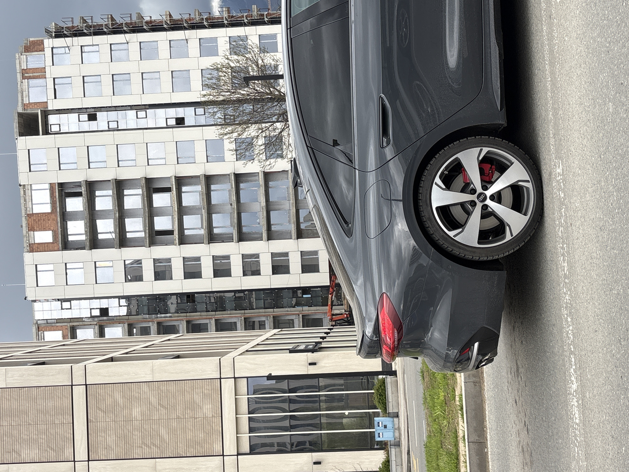 The image shows a parked gray Audi car on a street in Sofia, Bulgaria. Behind the car are two buildings. The first is a multi-story building, possibly under construction or renovation, with a grid-like pattern of windows and beige and white facade. The second is a modern building with a light beige or tan exterior and a horizontal pattern. The street appears paved, and a patch of green grass is seen adjacent to the curb. There are no visible people.
