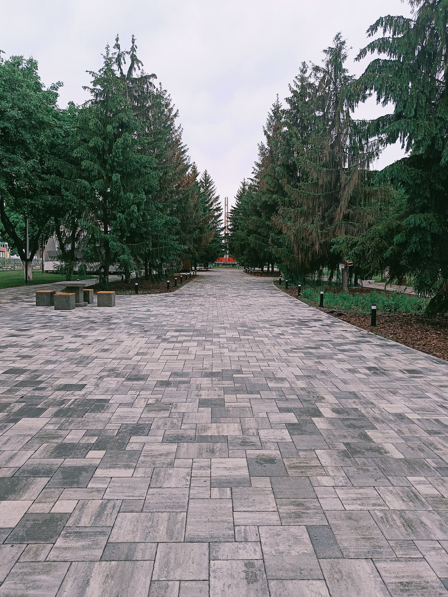 This is a ground-level, eye-level, medium shot of a paved pathway in a park in Gubkin, Russia. The pathway is made of large, rectangular stone pavers in varying shades of grey and light brown, arranged in a randomized pattern. The pathway curves gently to the right and extends into the distance, lined on both sides by tall evergreen trees.
On the left side of the pathway, closer to the foreground, there are several low, grey, concrete benches with wooden seating. In the middle ground on the left, there is a dome-shaped structure partially obscured by trees, and beyond that, a playground area with colorful structures.
On the right side of the pathway, in the middle ground, there are several modern, black, bollard-style lights spaced evenly apart, illuminating the path. Beyond the lights, there is a flowerbed with mulch and some greenery.
The trees are dense and dark green, with some of them appearing older and thinner. The sky above is overcast and light grey. In the far distance, beyond the trees, glimpses of red structures, possibly part of an urban landscape or an art installation, are visible. The overall atmosphere of the image is serene and naturalistic, despite the urban park setting.