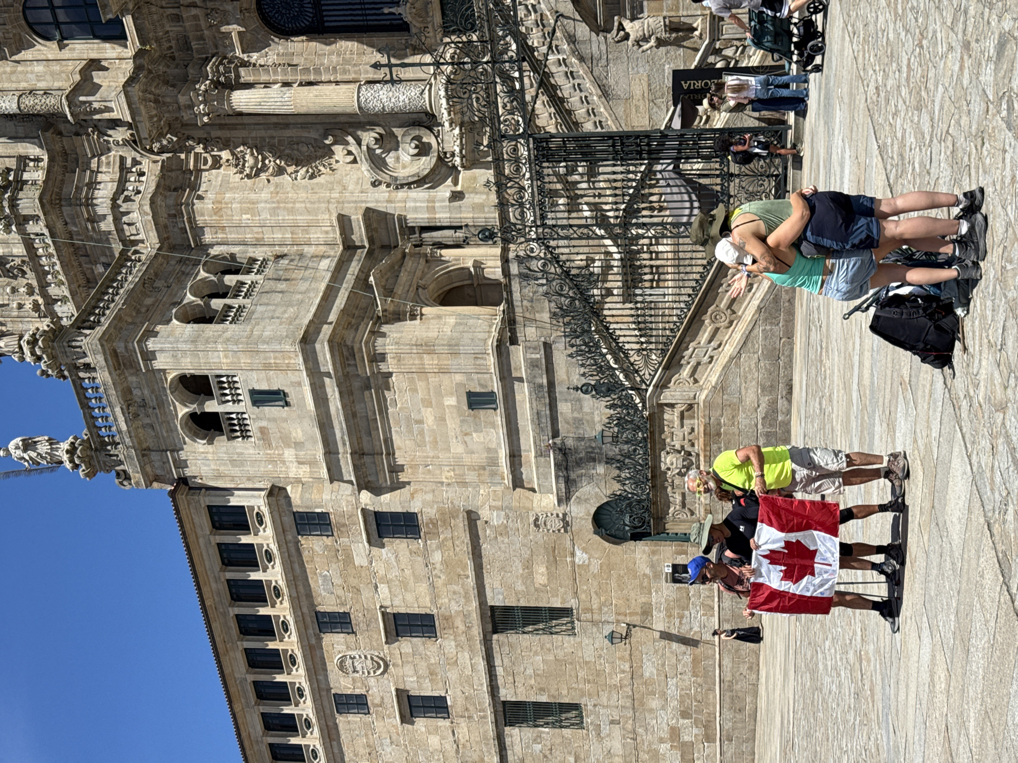 This media file captures a sunny day scene in Santiago de Compostela, Spain, focusing on people interacting with a historic building. The architecture is ornate and made of light-colored stone, featuring intricate carvings, columns, and numerous windows. A clear blue sky suggests good weather and likely mid-day or afternoon light, casting distinct shadows.
In the foreground, a group of people are visible. One couple is in an embrace, the woman wearing a green top and the man a blue tank top. They are standing on a paved area in front of the building. Further back, two men are holding a Canadian flag. One man wears a neon yellow shirt and sunglasses, while the other wears a hat and a blue cap. A black backpack is on the ground near the embracing couple. Other individuals and a stroller are also present in the background, suggesting a public space with tourist activity.
The scene appears to be a moment of leisure and sightseeing. The embracing couple might be experiencing a romantic moment, while the individuals with the flag could be tourists commemorating their visit. The overall emotion conveyed is one of enjoyment and exploration in a significant historical location. No specific text is clearly legible in the image, although a sign is visible on the building in the background.