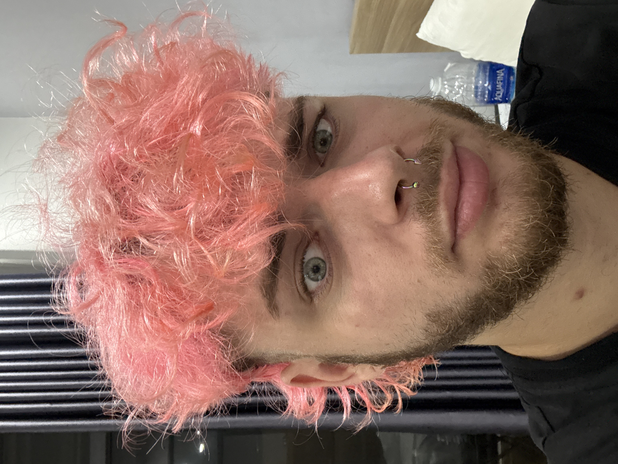 A man with pink curly hair, a beard, and nose piercings is shown in a close-up headshot. He has light eyes and fair skin. A water bottle is visible in the background along with a white pillow and a wooden headboard. The background suggests an indoor setting, possibly a hotel room. There is no indication in the image that it was taken in Đà Nẵng, Vietnam.