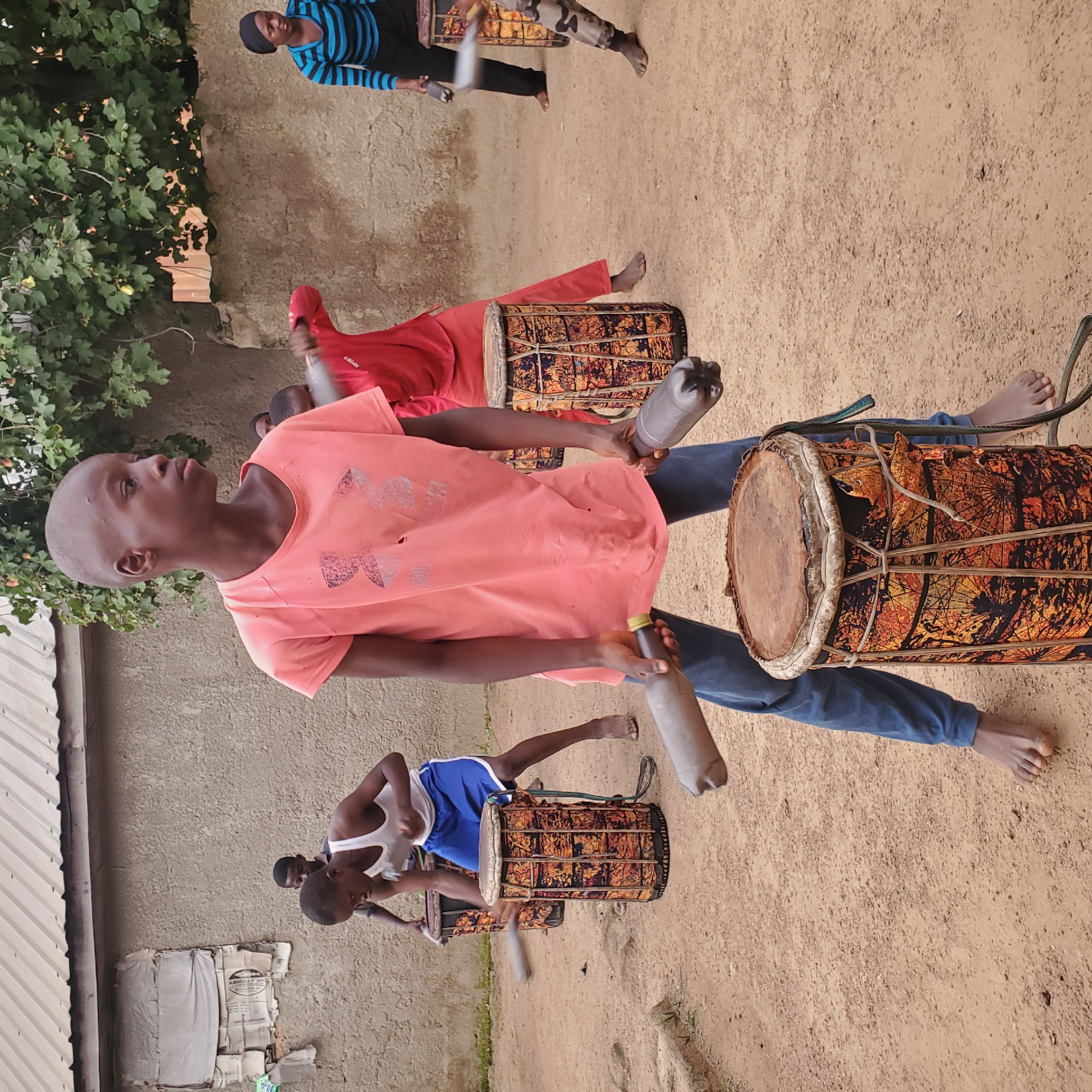 In an outdoor dirt courtyard, a group of children and one adult are gathered, some playing drums. A young boy in the foreground, wearing a coral t-shirt and blue jeans, stands with his legs apart and holds two cylindrical drumsticks made from plastic bottles, poised to strike a tall, patterned drum. He has a serious expression and is barefoot. Behind him, another boy in a white tank top and blue shorts is also playing a similar drum. Further back, a child in a red long-sleeved shirt and pants is actively drumming, and another person in a striped blue and black top and black pants is visible to the right, also holding a drumstick. The environment features a rough, textured wall behind the group and a corrugated metal roof structure above. Lush green foliage from a tree is visible in the upper right quadrant of the image. The ground is dry and dusty.