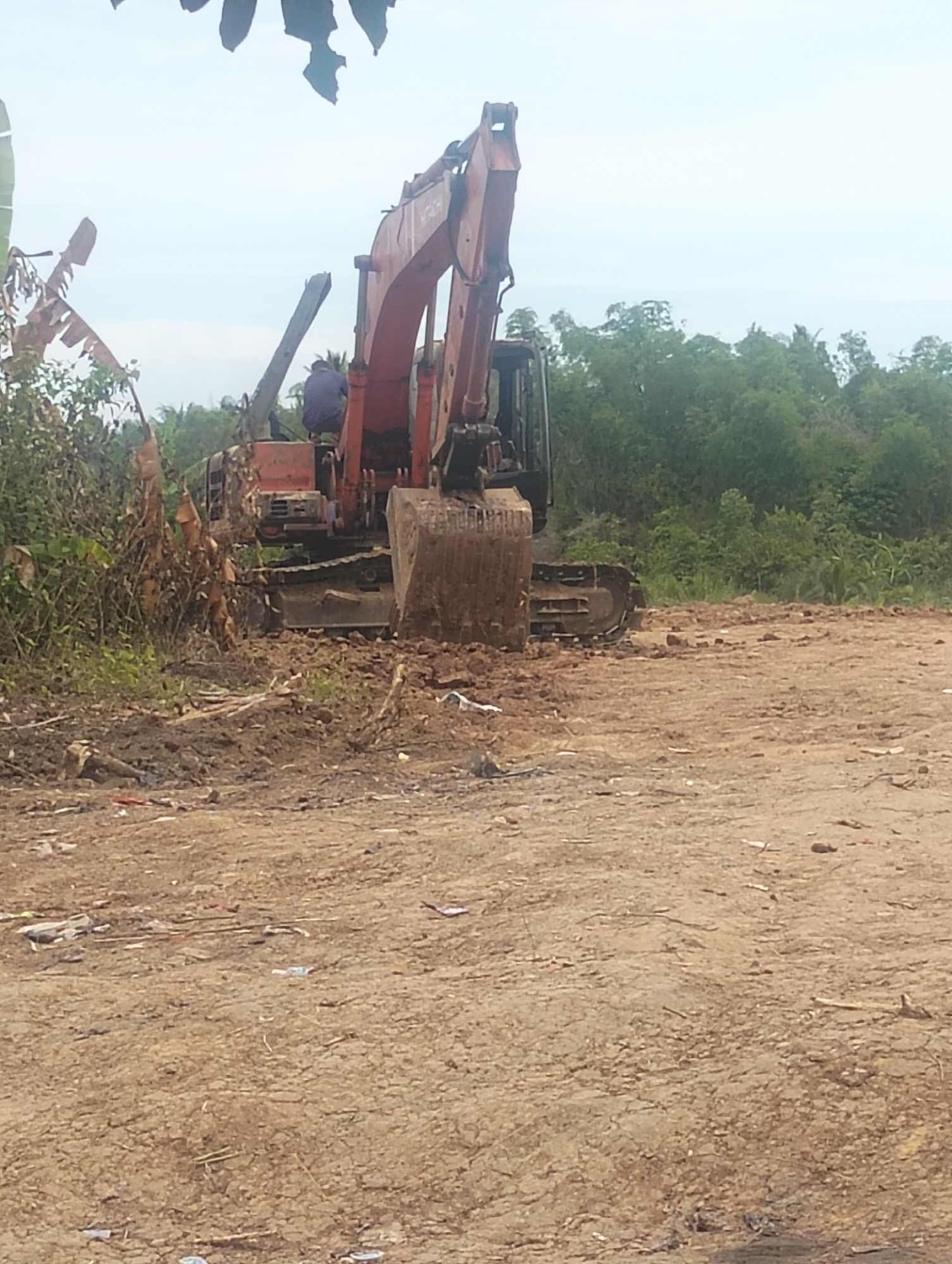 An excavator is parked on a dirt lot in a rural area, possibly in Payagaboh, Indonesia, as indicated by the presence of tropical vegetation like banana trees. The excavator, a large orange and black machine, appears to be idle, with a single person, wearing a dark blue shirt and possibly dark pants, seated in the operator's cabin. The ground is uneven and covered with loose dirt, rocks, and debris, suggesting an ongoing construction or excavation project, or possibly a waste disposal site due to scattered plastic waste. In the background, a dense line of green trees forms the horizon under a hazy, overcast sky. The overall scene depicts a work-in-progress or a site that has recently undergone significant earthmoving.