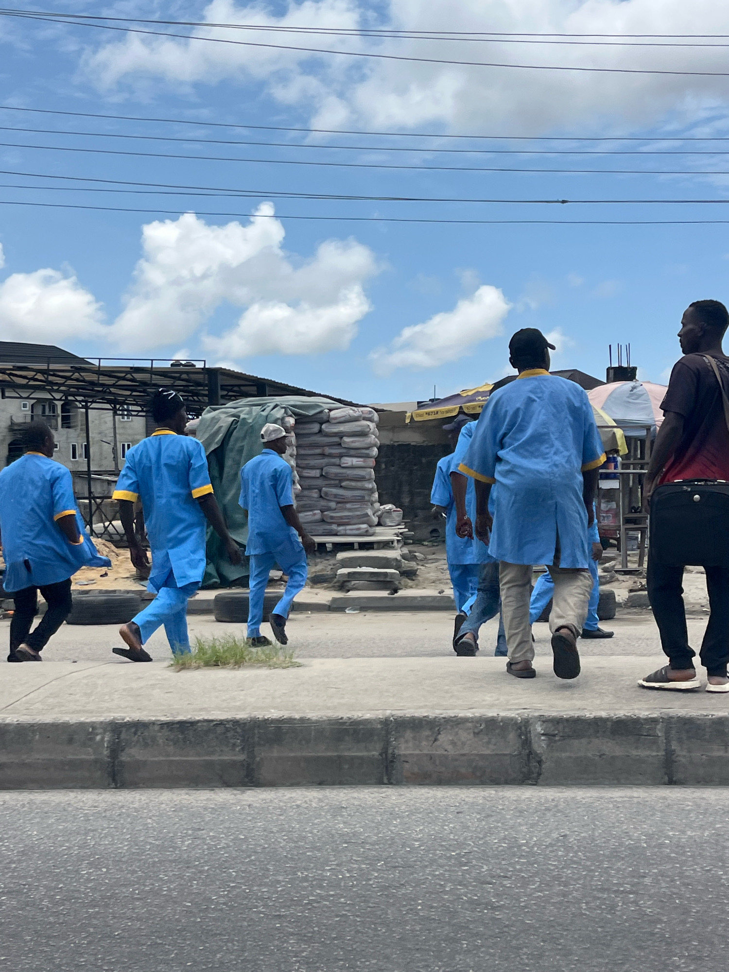 A group of men, most wearing blue uniforms with yellow trim, walk along a sidewalk in Ado, Nigeria. The men are walking in the same direction, away from the camera, and appear to be engaged in some sort of work or labor. The environment is urban, with a paved road in the foreground and buildings and a stack of bagged goods in the background. The sky is blue with scattered white clouds. One man on the right of the image wears a dark shirt and pants and carries a bag. Another man in the foreground on the left wears a dark shirt and pants and has a blue garment draped over his shoulder. The men in blue uniforms are likely workers, possibly construction or logistics personnel, given the context of the bagged materials.
