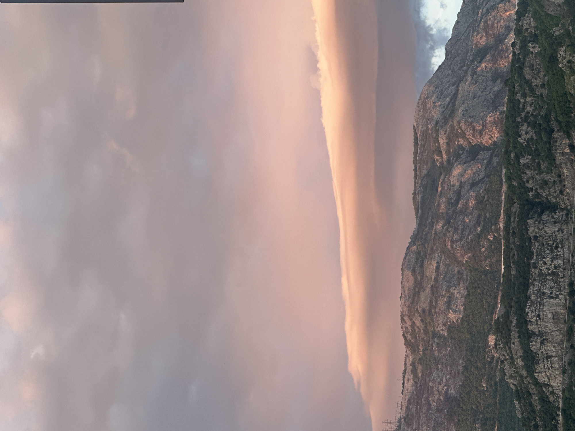This image captures a majestic natural landscape, likely at either sunrise or sunset, near Budva, Montenegro. A towering, rugged mountain range dominates the right and lower portions of the frame. Its craggy, grey and brown rock faces are illuminated by warm, low-angle light, highlighting textured surfaces and showing hints of pink and orange. Patches of dark green vegetation cling to the mountain slopes, particularly denser on the lower right side.
The sky, filling the left and upper parts of the image, presents a soft palette of light pink, subtle orange, and pale grey clouds. A striking and unusually elongated cloud formation, possibly a lenticular cloud, stretches horizontally across the sky, glowing brightly with the warm light, creating a strong visual line against the more diffuse background clouds.
The scene exudes tranquility and natural grandeur, with no explicit human activity visible. The warm lighting suggests either early morning or late afternoon. The mountainous terrain is consistent with the geographical features of Montenegro, where the Dinaric Alps meet the Adriatic coast. There is no visible text in the image. Faint structures resembling power lines or poles can be vaguely discerned at the base of the mountains on the far lower left, indicating some human infrastructure in the expansive natural setting.
