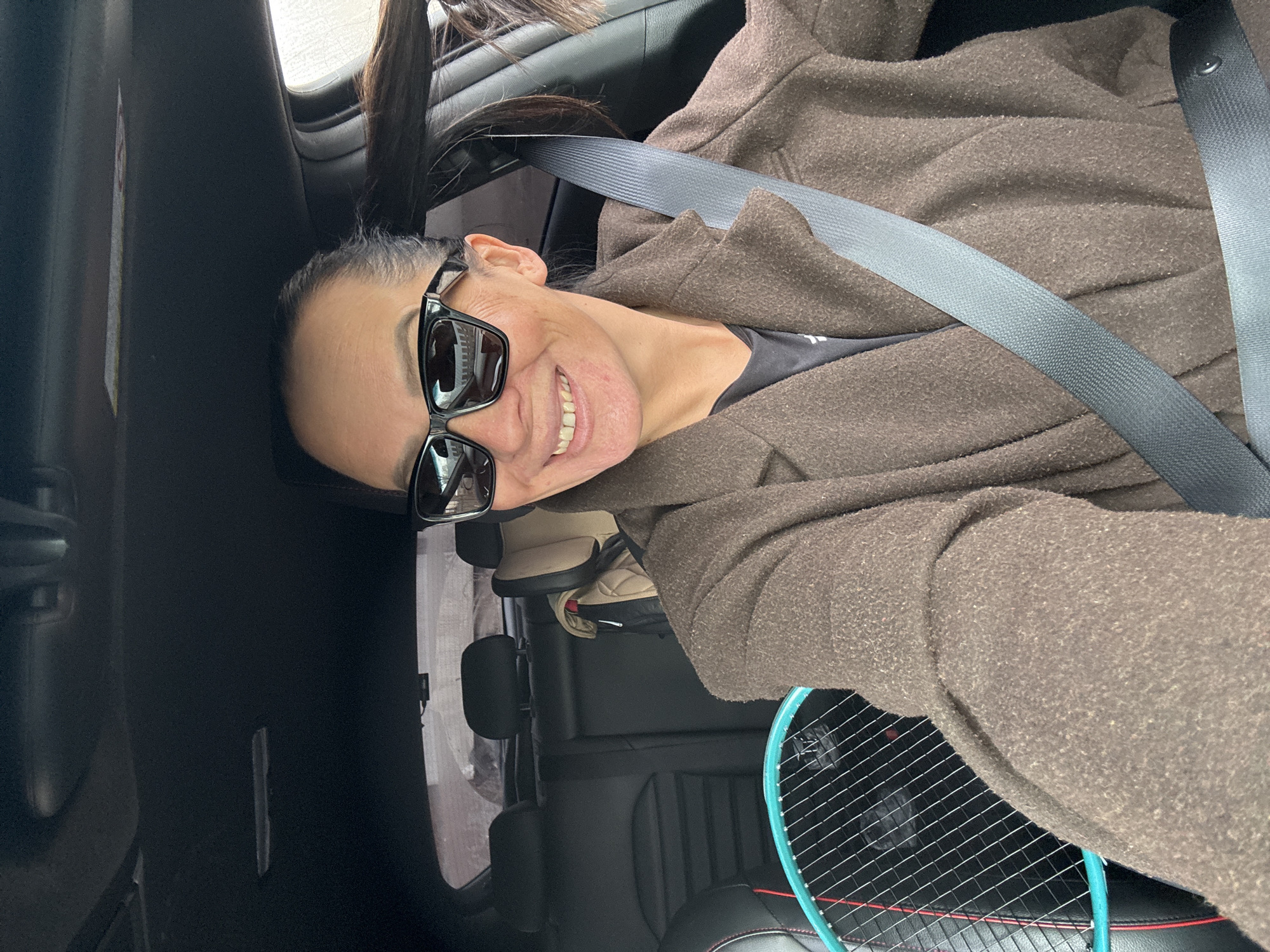 The image shows a person sitting in a car, wearing sunglasses and a brown coat, secured with a seatbelt. A tennis racket is visible in the back seat. The location in Chaikovsky, Russia, is not determinable from the provided visual information.