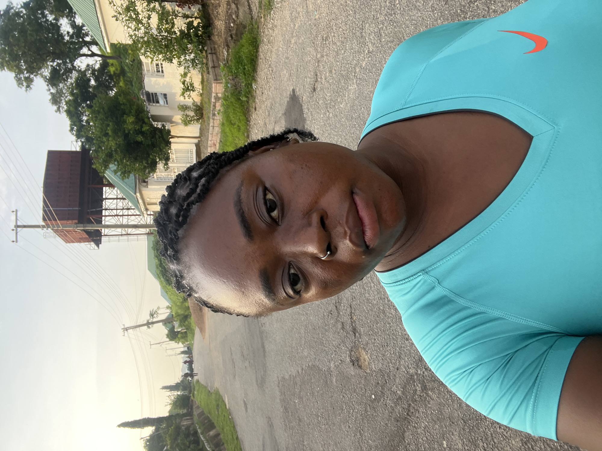 A medium shot, selfie-style photograph features a Black woman with dark braided hair and a nose ring. She is wearing a turquoise v-neck t-shirt with a red Nike swoosh logo on the right chest. She is standing on a paved road in what appears to be a residential area. Behind her, the road stretches into the distance, lined with grass, vegetation, and houses with green roofs. In the background, a large, dark red water tower stands on a metal frame. Power lines crisscross the sky. The sky is overcast, suggesting it may be early morning or late afternoon. The overall scene depicts a serene, outdoor setting, possibly a neighborhood in Abuja, Nigeria, given the characteristic architecture and infrastructure.