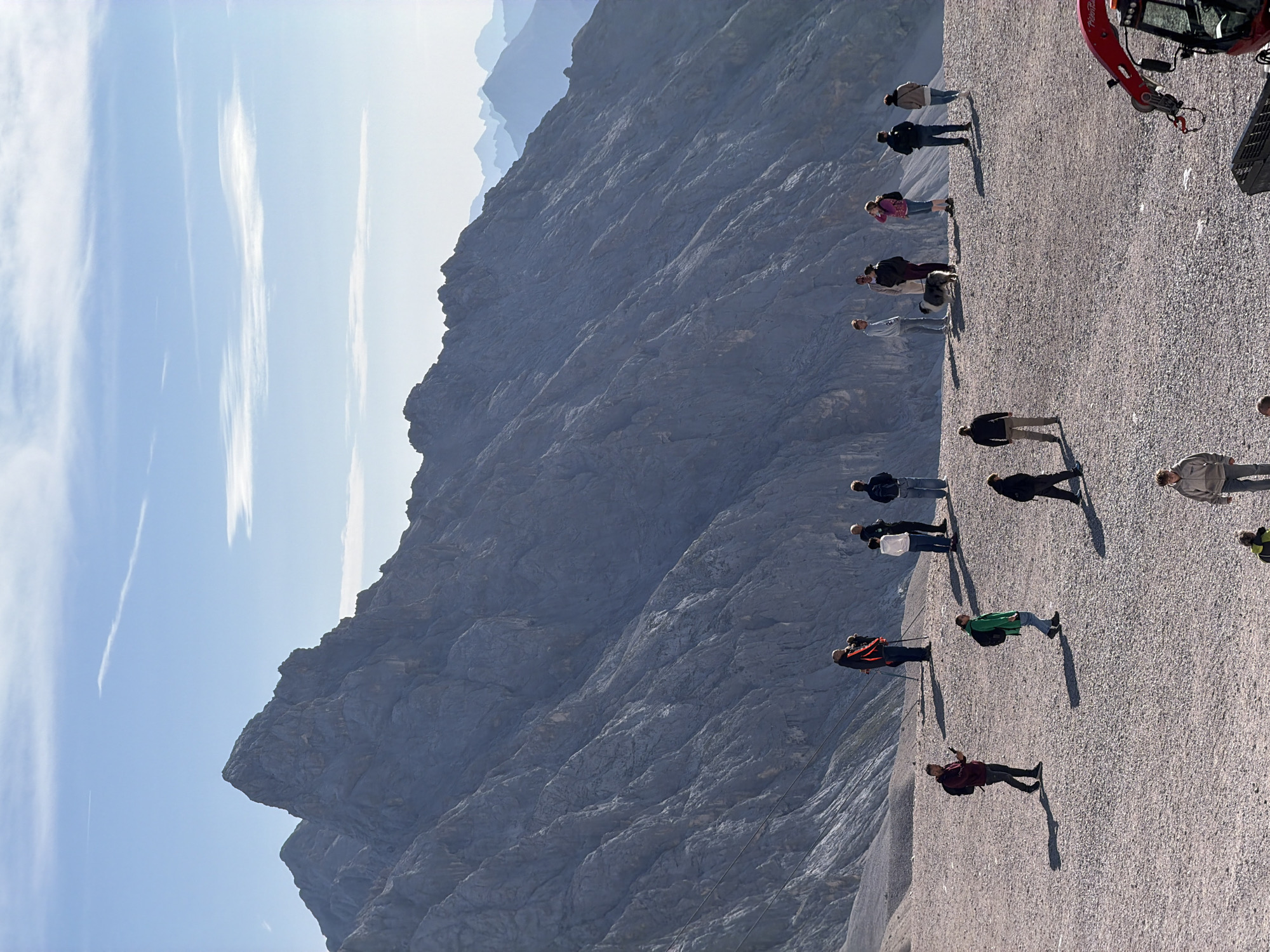 In Garmisch-Partenkirchen, Germany, atop a rocky, gravel-covered mountain plateau, a group of individuals are gathered against a backdrop of imposing, gray, jagged mountain peaks under a clear blue sky with wispy white clouds. The people are dispersed across the foreground and midground, engaged in various activities. Some are standing and observing the scenery, while others appear to be walking. One person in the mid-left is holding a phone. A person in the mid-right is accompanied by a fluffy, light-colored dog. In the far right of the image, a red construction vehicle with a robotic arm is partially visible. The overall scene suggests a popular tourist destination offering panoramic mountain views.