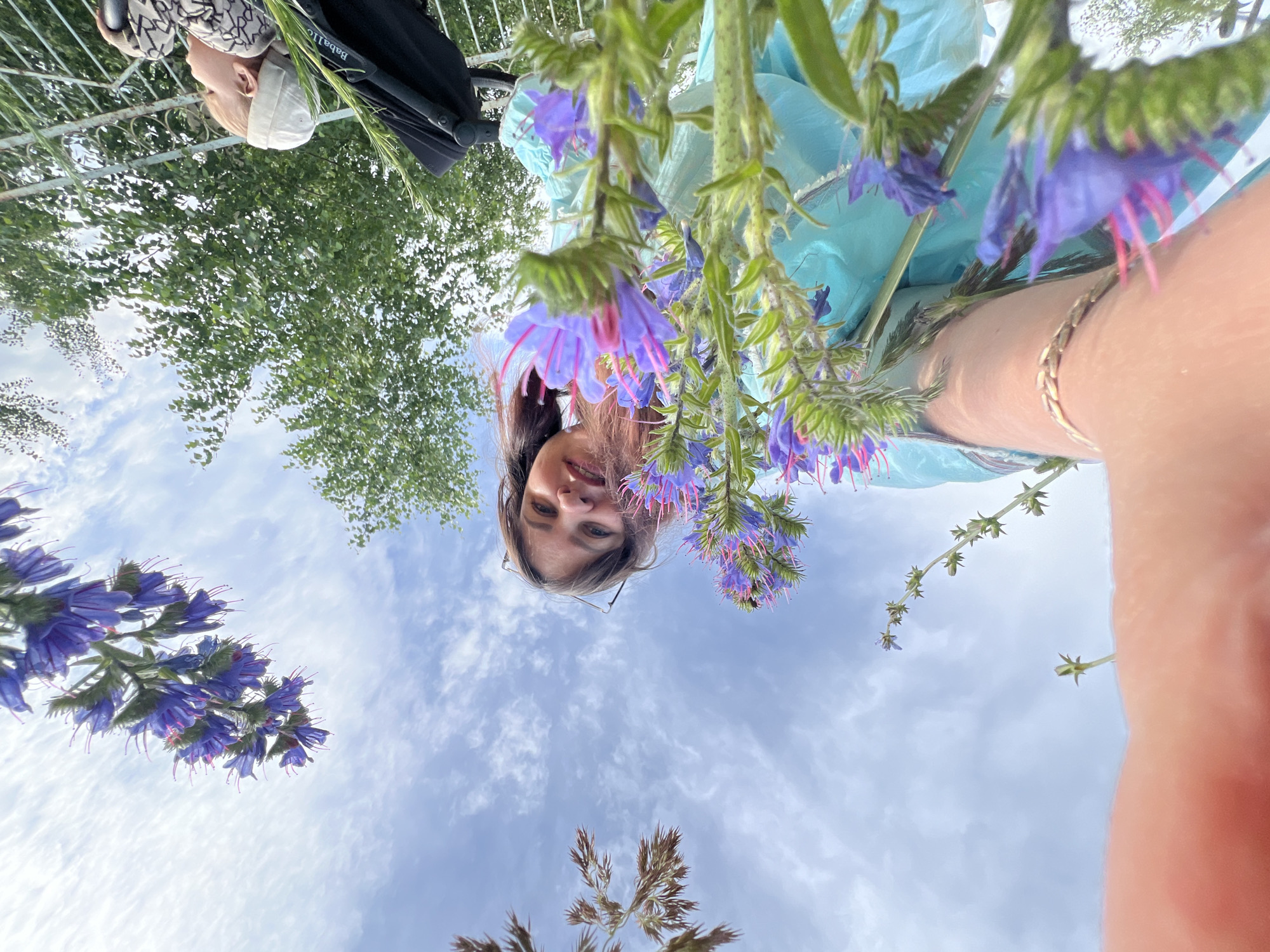 This is a selfie taken from a low angle, looking up at the sky, with flowers in the foreground. The image features a young woman with brown hair and glasses, smiling at the camera. She is wearing a light blue garment and a silver bracelet on her arm. Behind her, purple flowers with pink stamens are in bloom, with green stems and leaves. The background consists of a bright blue sky with scattered white clouds, indicating a clear and sunny day. Trees are visible in the upper left and right corners of the image.
In the upper left corner of the image, partially obscured by greenery and a stroller, a baby is visible wearing a white hat. A black stroller is also present.
The image appears to be taken outdoors, likely in a garden or park setting in Noviy, Russia, during daylight hours. The overall mood is cheerful and relaxed.