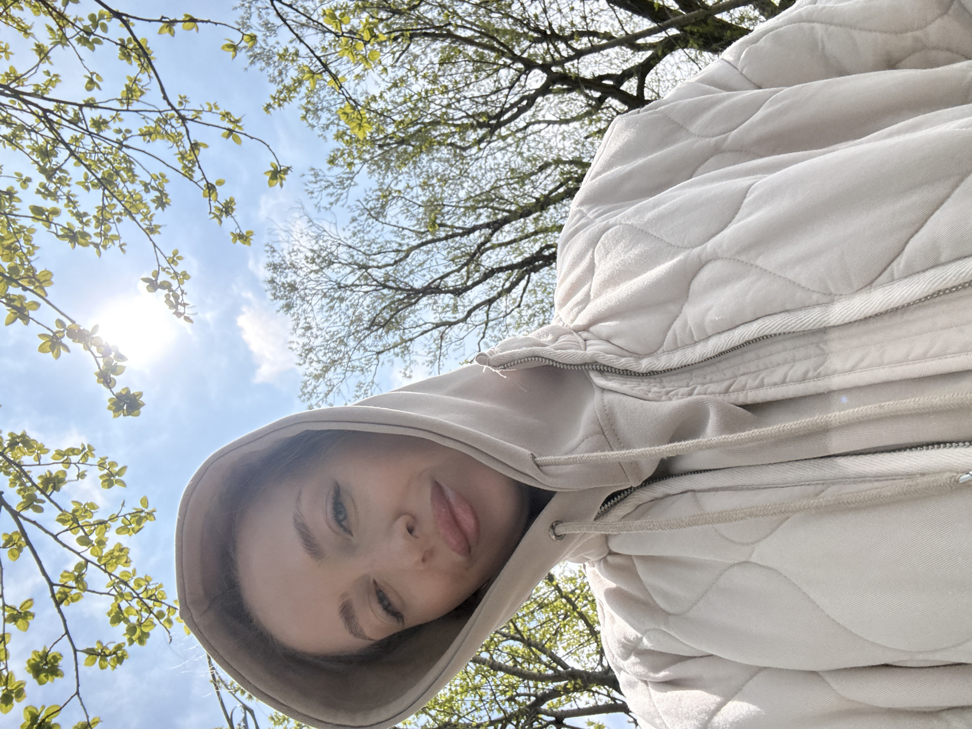 This low-angle outdoor portrait features a young woman looking directly at the camera, taken in what appears to be a park or natural setting in Petropavlovsk-Kamchatsky, Russia. Her face, primarily the left side, and a portion of her upper body are visible. She wears a light-colored, possibly beige or off-white, quilted hooded jacket with a visible zipper and drawstrings. Her expression is playful, with her tongue slightly out. Her eyes are open and focused on the viewer.
The background is dominated by a bright blue sky with scattered white clouds. The sun is partially visible in the upper left, casting a bright light and creating a lens flare. Silhouetted against the sky are numerous tree branches adorned with vibrant, fresh green leaves, indicating springtime or early summer. The perspective suggests the photo was taken from a low vantage point, looking up through the foliage. The scene conveys a bright, sunny daytime environment. There is no visible text in the image.