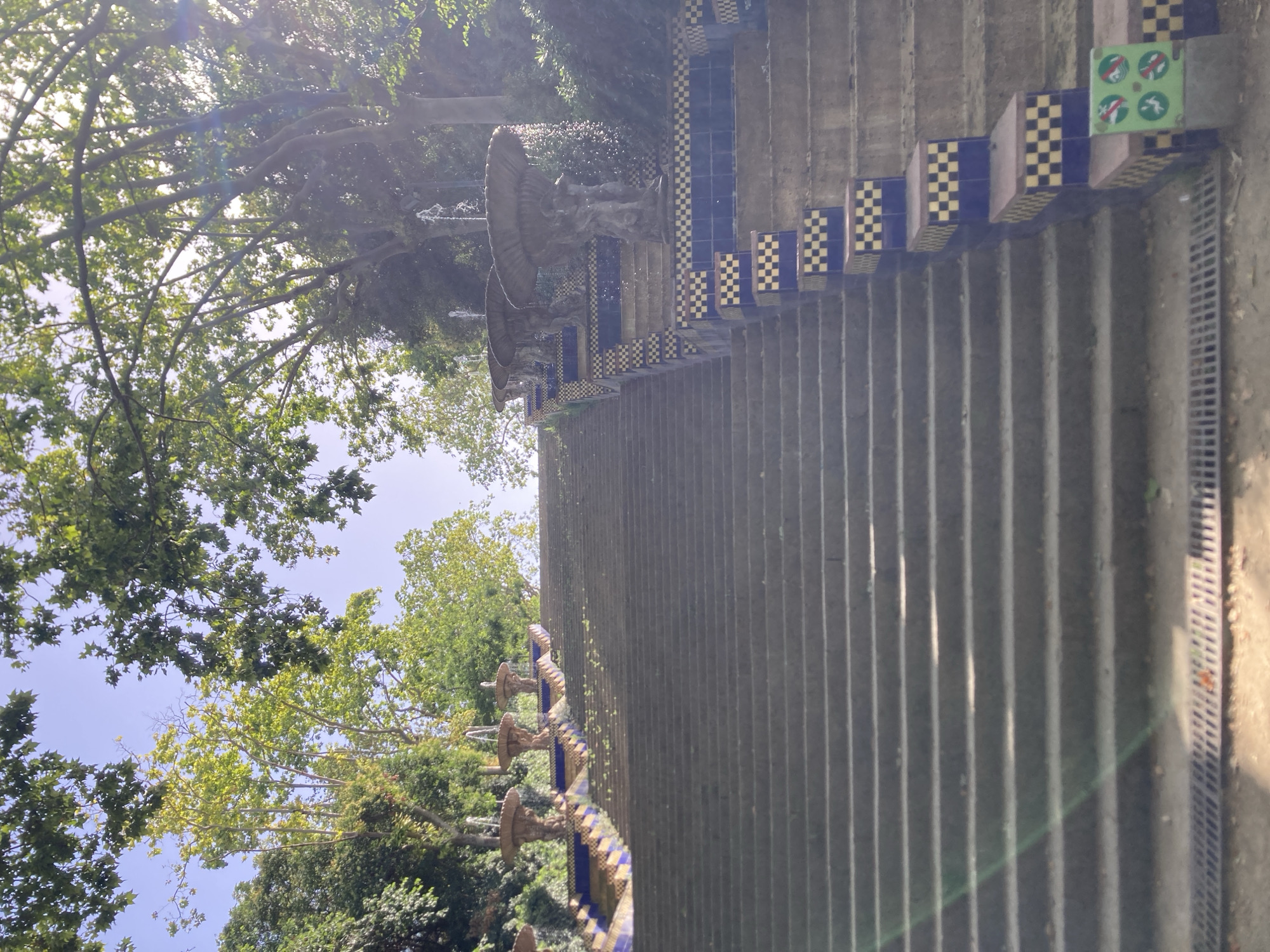 The image depicts a wide, gray stone staircase ascending a hillside, lined with decorative tiled risers and fountains. Lush green trees with dappled sunlight filter through the canopy, suggesting an outdoor park setting. At the top of the stairs, several tiered fountains with water spouting upward are visible. The tiled risers feature a pattern of blue and yellow squares. The overall impression is one of a serene, artistic public space, likely within Barcelona, Spain, given the architectural style often found in the city's parks and gardens. The scene is devoid of people, and the lighting indicates it is daytime.