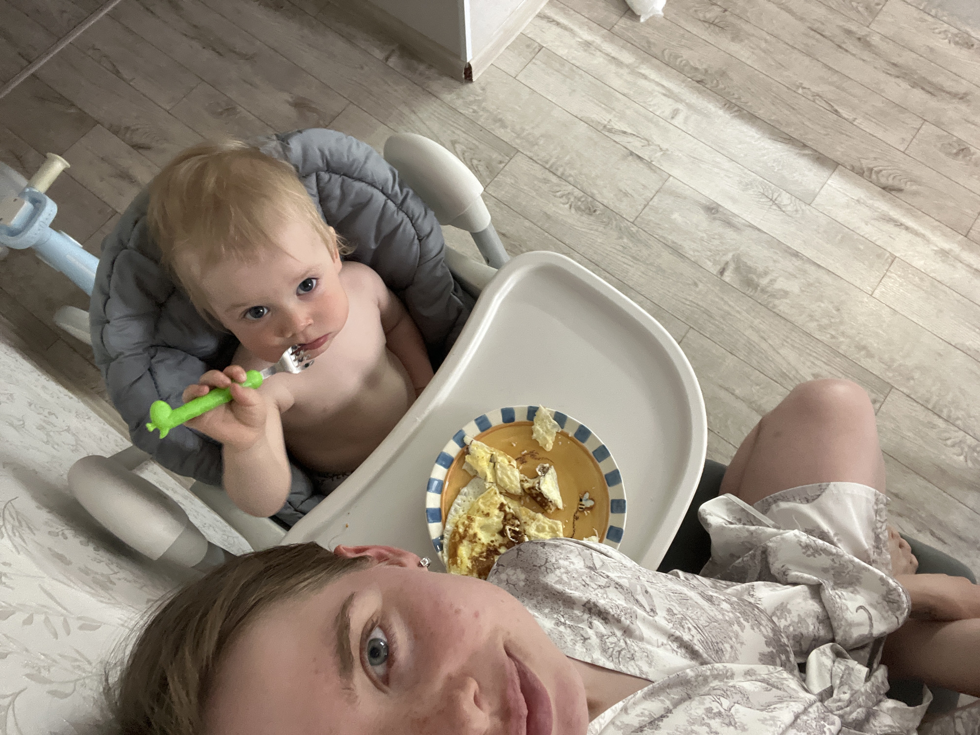 A top-down, high-angle interior photograph taken in Chaikovsky, Russia, depicts an adult female and an infant during a mealtime. In the left foreground, the upper left portion of a light-skinned adult female's face is visible, showing one blue eye, freckles on her cheek, and short brown hair. She is wearing a white robe with a grey and white patterned design. Her right arm and leg are partially visible at the bottom.
To her right, a light-skinned infant with blonde hair and blue eyes is seated in a white high chair with a grey padded insert, looking directly at the camera. The infant appears bare-chested. In the infant's right hand, a green, dinosaur-shaped fork with food particles on its tines is held.
On the white high chair tray, a round plate with a blue and white striped rim, a yellow center, and a bee graphic holds a portion of chopped, cooked eggs, some pieces exhibiting slight browning.
Behind the high chair, a light blue children's ride-on toy with white handlebars is partially visible. The wall in the background is covered with light-toned patterned wallpaper. The floor is covered with light-colored wood-look laminate planks.