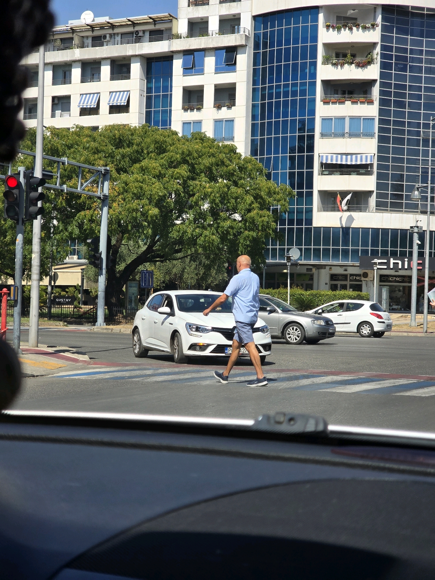 The image depicts a street scene in Podgorica, Montenegro, likely on a sunny day given the bright lighting and clear sky. In the foreground, the interior of a vehicle is visible, suggesting the photograph was taken from a car. A pedestrian crossing is in the middle ground, with a bald man in a blue shirt and shorts walking across it. He appears to be reaching out towards a white car that is stopped at the crossing. To the left of the pedestrian, a traffic light is illuminated red. Behind the pedestrian and the cars, a modern building with blue glass windows dominates the background, interspersed with trees and other urban structures. The overall environment is a busy city intersection with traffic and pedestrian activity.