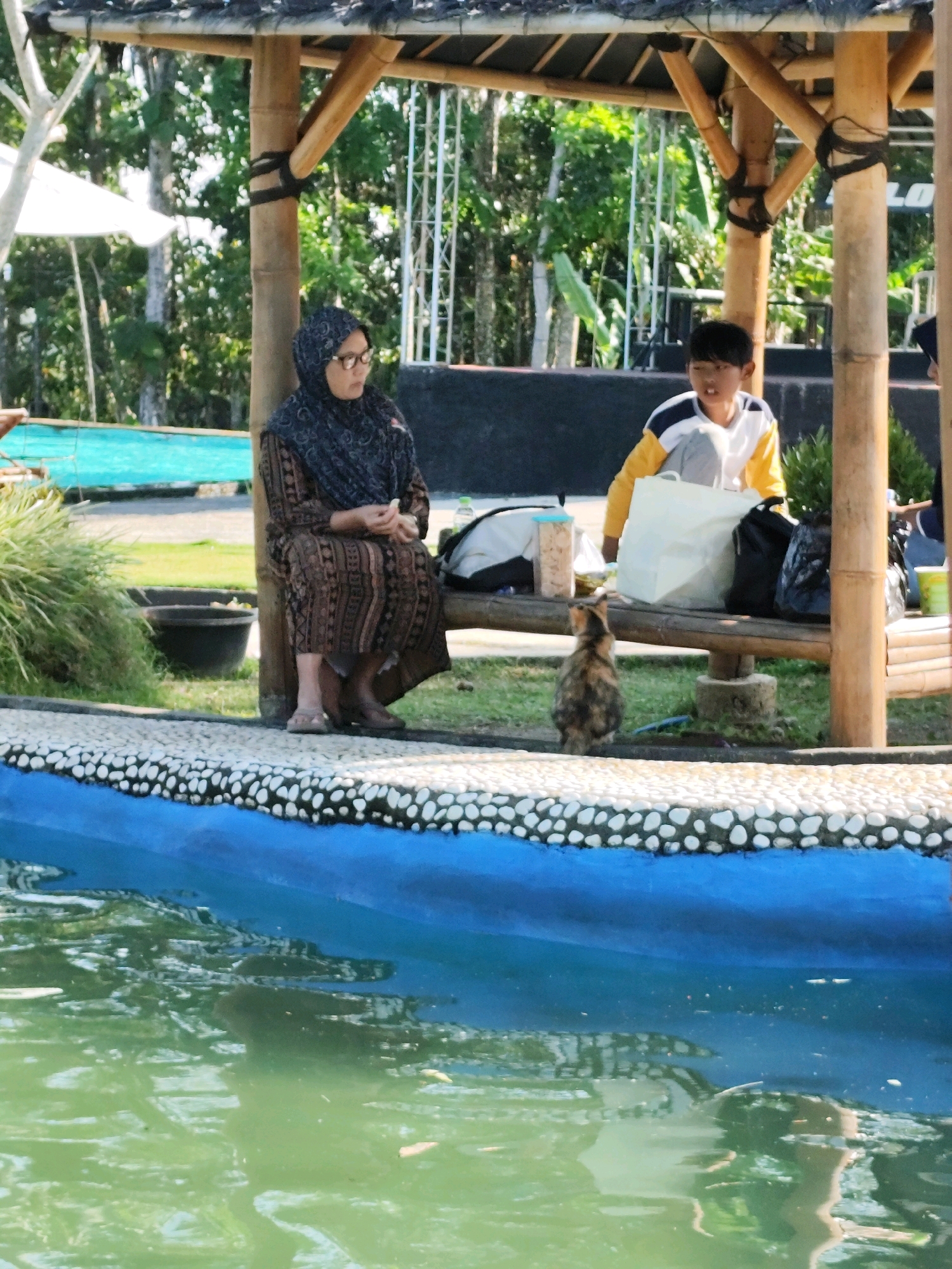This image depicts an outdoor scene at what appears to be a recreational area, possibly a resort or a park with a pool, located in Unknown, Indonesia. The foreground features a portion of a swimming pool with green, reflective water. The pool is bordered by a textured edge with white and grey pebbles. In the middle ground, a bamboo-constructed gazebo or shelter provides shade. A woman wearing a patterned headscarf and a brown, patterned dress is seated on a wooden bench within the shelter, looking towards the viewer. Next to her, a young boy wearing a yellow and white long-sleeved shirt and grey pants is also seated. A calico cat is positioned on the pebbled border, facing the boy and looking upwards. Several bags and containers are placed on the bench. In the background, lush green trees and vegetation are visible, along with elements of the gazebo's structure.