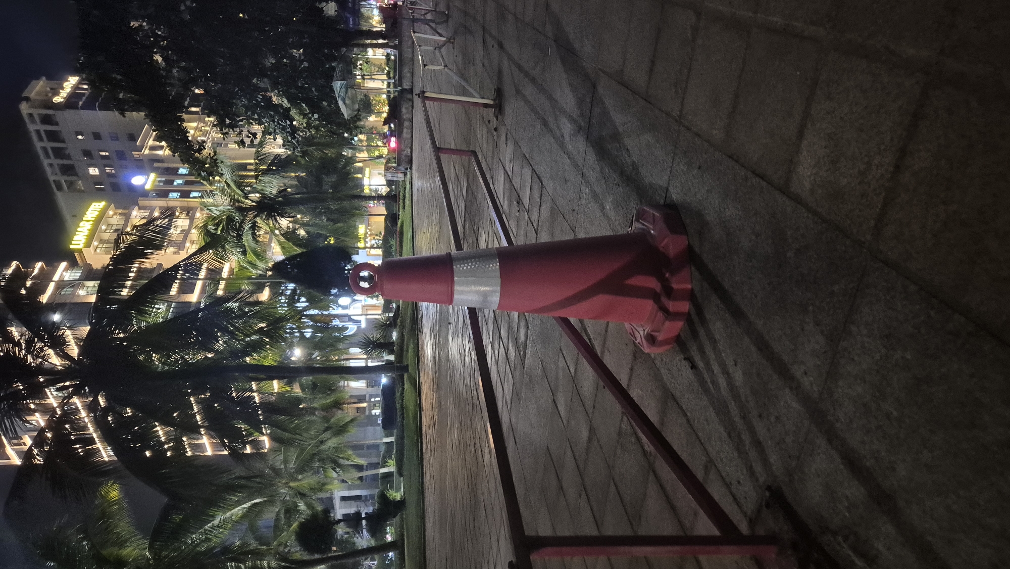 A red traffic cone with a reflective silver band is positioned on a paved walkway at night. The cone is in the foreground, slightly off-center to the right, casting a long shadow. Behind the cone, a red metal railing runs horizontally across the image. In the background, a modern hotel, identified as the "LUNAR HOTEL" by its illuminated sign, stands tall with many windows lit up. Several palm trees are scattered throughout the scene, their fronds silhouetted against the building. The walkway is made of rectangular stone tiles. The overall setting appears to be an outdoor public area in Đà Nẵng, Vietnam, possibly a promenade or near a hotel entrance.
