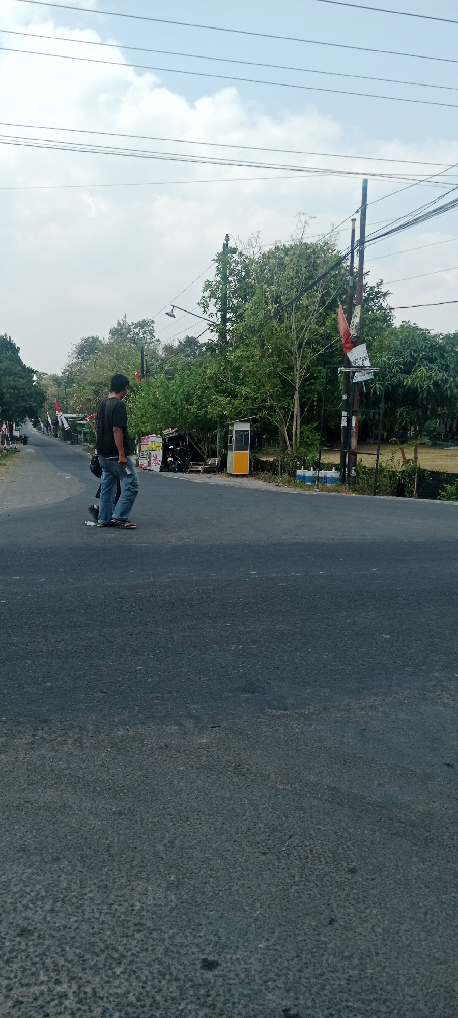 A street scene in Maguwoharjo, Indonesia, captures two individuals crossing a paved road under an overcast sky. The road appears to be a rural or semi-rural intersection with trees and vegetation lining the sides. A small, yellow and white structure, possibly a roadside kiosk or small shop, is visible on the right. Several motorcycles are parked near a shaded area. Power lines crisscross the sky. One person, wearing a dark t-shirt and blue jeans, is mid-stride. The second person, partially obscured, is walking alongside. Flags, seemingly red and white, are displayed on poles along the roadside, suggesting a local celebration or national pride. The overall impression is a snapshot of everyday life in a community.