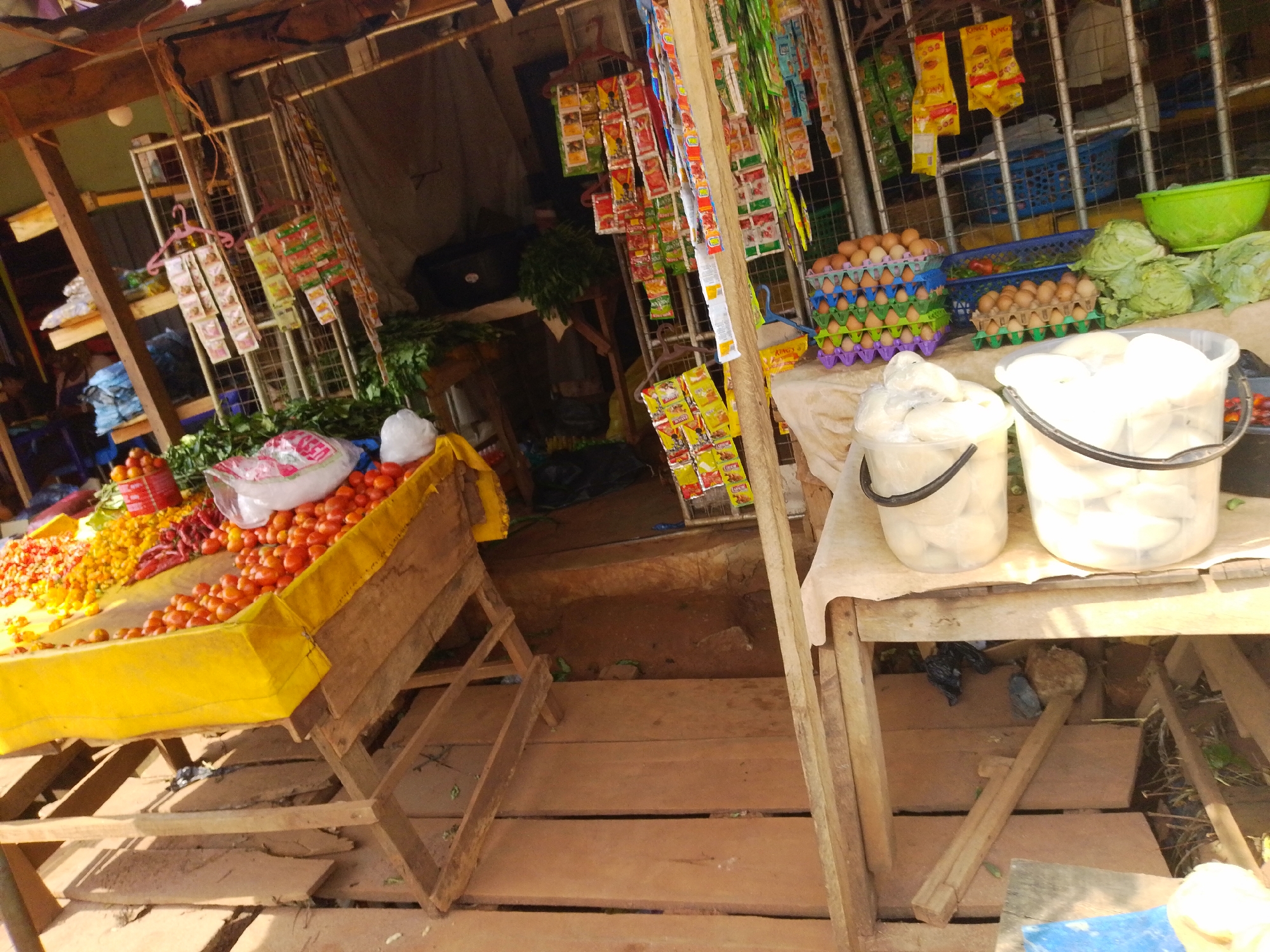 This is a market stall in Nsukka, Nigeria, with produce and other goods for sale. The stall appears to be made of wood and is set up outdoors. In the foreground, there are two large plastic buckets filled with what appear to be yams or other root vegetables, wrapped in plastic. To the left, a yellow-covered table displays a variety of fresh produce, including tomatoes, peppers, and other colorful vegetables. In the background, there are racks of packaged goods, likely food items, hanging from the stall. Eggs are also visible in colorful plastic trays. The ground is covered with wooden planks, and the overall impression is that of a bustling, everyday market scene.