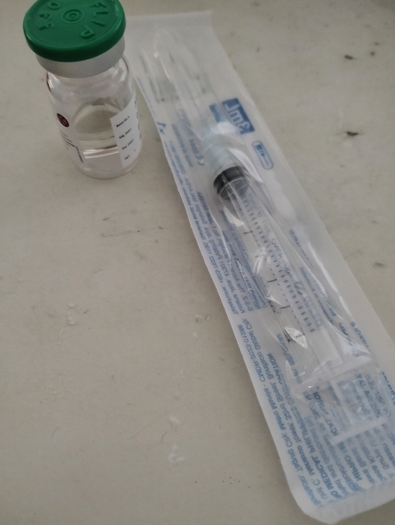 The image shows a medical scene, likely in a private home or clinic, as indicated by the presence of a vial of medicine and a syringe. The vial is clear glass with a green cap, and it appears to contain a clear liquid. The syringe is a standard disposable plastic syringe with a needle, packaged in a clear plastic wrapper. The background is a plain, light-colored surface, possibly a table or countertop. There is no indication of the location in Malang, Indonesia. The image is a close-up shot, and the lighting is natural and diffused.