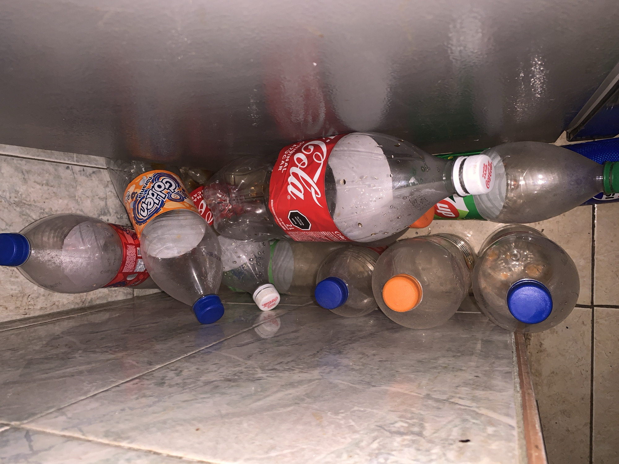 The image displays a collection of numerous used plastic beverage bottles, predominantly clear PET plastic, arranged horizontally on a tiled floor beneath a reflective metallic surface. The viewpoint is from a low angle, looking upwards at the bottles.
Prominent among the bottles is a Coca-Cola bottle featuring its iconic red label, which clearly shows the "Coca-Cola" logo, "ORIGINAL SABOR" (Original Flavor), and a warning label indicating "ALTO EN AZÚCARES" (High in Sugars). This bottle is topped with a white cap. Adjacent to it, another bottle displays an orange label with the brand "Golden" in large blue lettering, likely "Golden Naranja" (Orange) flavor, and has a blue cap. A partially visible bottle with a green label and a white cap appears to be a "7 Up" product. Other clear plastic bottles with various colored caps, including blue, orange, and green, are also scattered throughout the collection. Many bottles exhibit condensation or residual droplets, suggesting they once held liquids.
The bottles are tightly packed together, creating a sense of a storage area, possibly for recycling. The floor is covered with light-colored, possibly grey or beige, tiles with distinct grout lines. The overhead metallic surface, possibly the underside of a shelf or appliance, reflects parts of the bottles and ambient light. The setting suggests an indoor utility space or a corner within a home in Santa Ana, Venezuela. There are no people visible, and no specific activity is taking place beyond the static arrangement of the bottles.