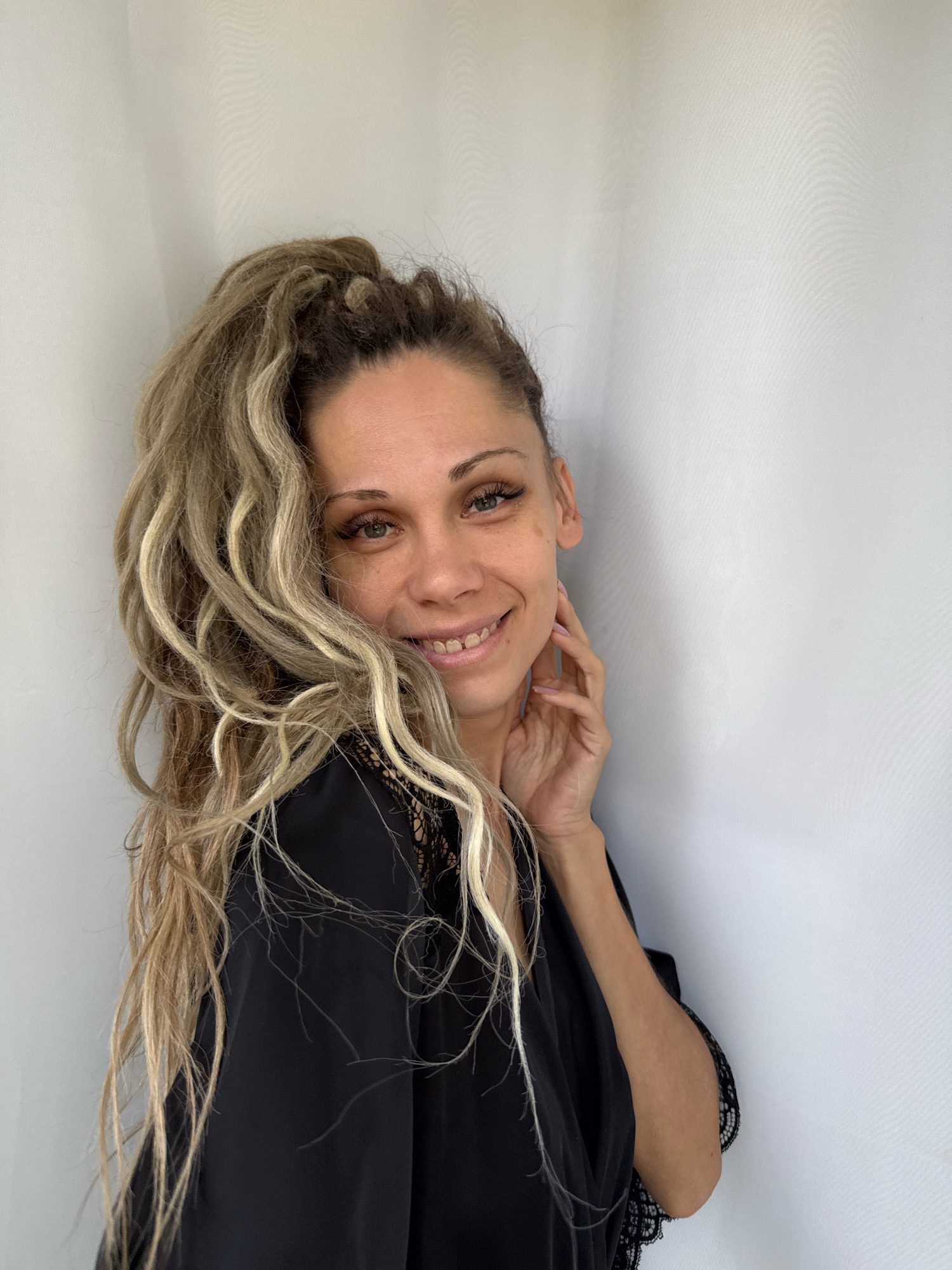 A woman with blonde dreadlocks pulled into a high ponytail smiles and touches her face. She is wearing a black robe with lace trim. The background is a plain white curtain. This image was not taken in Rusanovo, Russia.