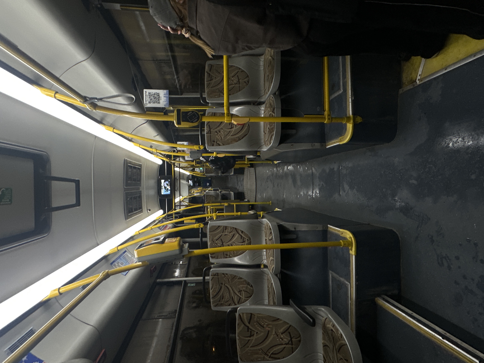 The image shows the interior of a public bus, likely in Minsk, Belarus, given the visible QR code with Russian text "ОПЛАТИ" which means "Pay" and a QR code for payment. The bus is mostly empty, with only one person visible on the right side of the image, a female wearing a dark jacket and a grey cap. She is facing away from the camera and appears to be about to exit or has just boarded. The seats are arranged in a typical bus configuration, with two seats on the left and two on the right side of the aisle. The seats are upholstered in a patterned fabric. Yellow handrails are installed throughout the bus for passengers to hold onto. The lighting inside the bus is from long fluorescent tubes on the ceiling. The floor of the bus appears wet, suggesting it may have recently rained or been cleaned. There are various signs and equipment visible inside the bus, including ticket validators and what appears to be a small screen displaying some form of information. The overall impression is of an ordinary public transportation setting.