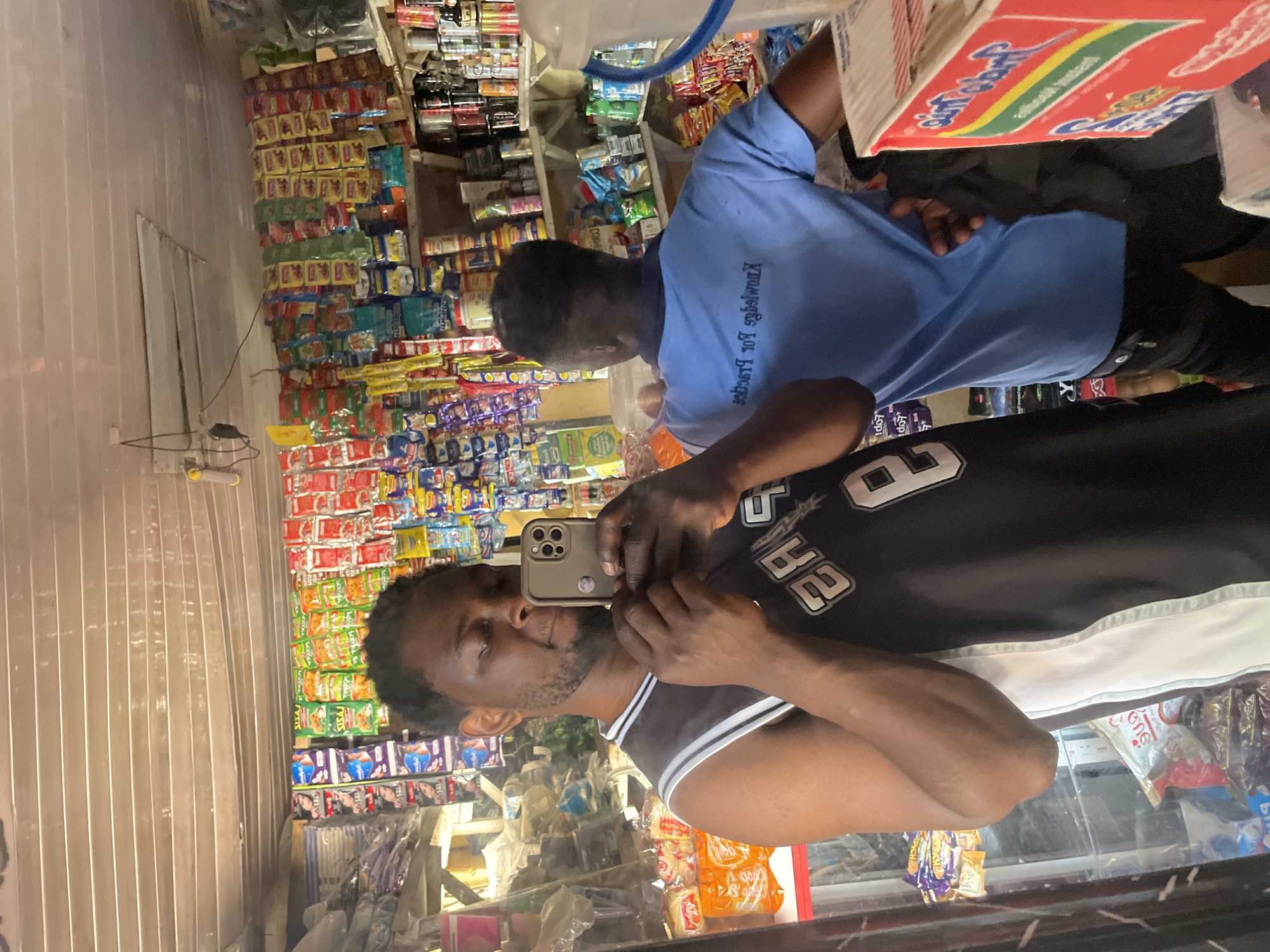 A man is taking a selfie in a convenience store in Durumi, Nigeria. He is wearing a black and white basketball jersey. Behind him, another man wearing a blue shirt is visible. The store is filled with shelves stocked with various food items, snacks, and drinks. The lighting in the store is bright, with fluorescent lights overhead. The shelves are organized and well-stocked, indicating a busy and successful business.