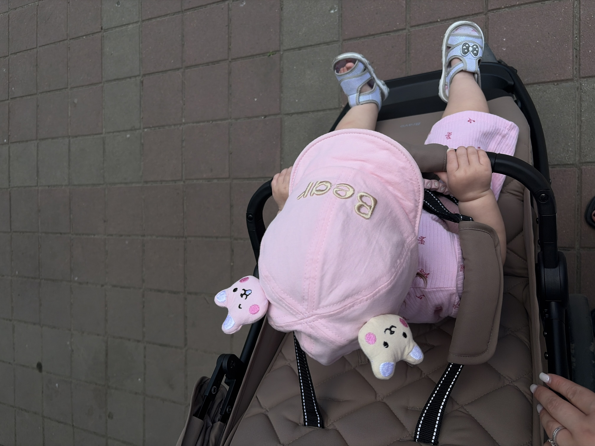 An overhead shot captures a toddler seated in a brown-quilted stroller on a paved surface in the city of Krasnodar, Russia. The child, viewed from above, wears a light pink short-sleeved top, light blue/grey open-toed sandals (the right sandal featuring a decorative bow), and a light pink bucket hat with "Bear" embroidered in gold script on the brim. Two small, plush toys are attached or resting on the hat: a pink rabbit-like figure with a winking face on top, and a cream-colored bear-like figure with pink polka-dot cheeks on the left side of the hat. The child's hands are clasped around the black bumper bar of the stroller. In the lower-left corner, an adult's left hand, featuring white-tipped nails and a silver ring on the ring finger, is visible, resting near the stroller's frame. The stroller itself has black frame components, and the word "Bravo" is subtly visible on one part of the frame. The ground beneath is comprised of alternating reddish-brown and grayish square paving stones, typical of an outdoor pedestrian area. The scene is evenly lit, indicating daylight.