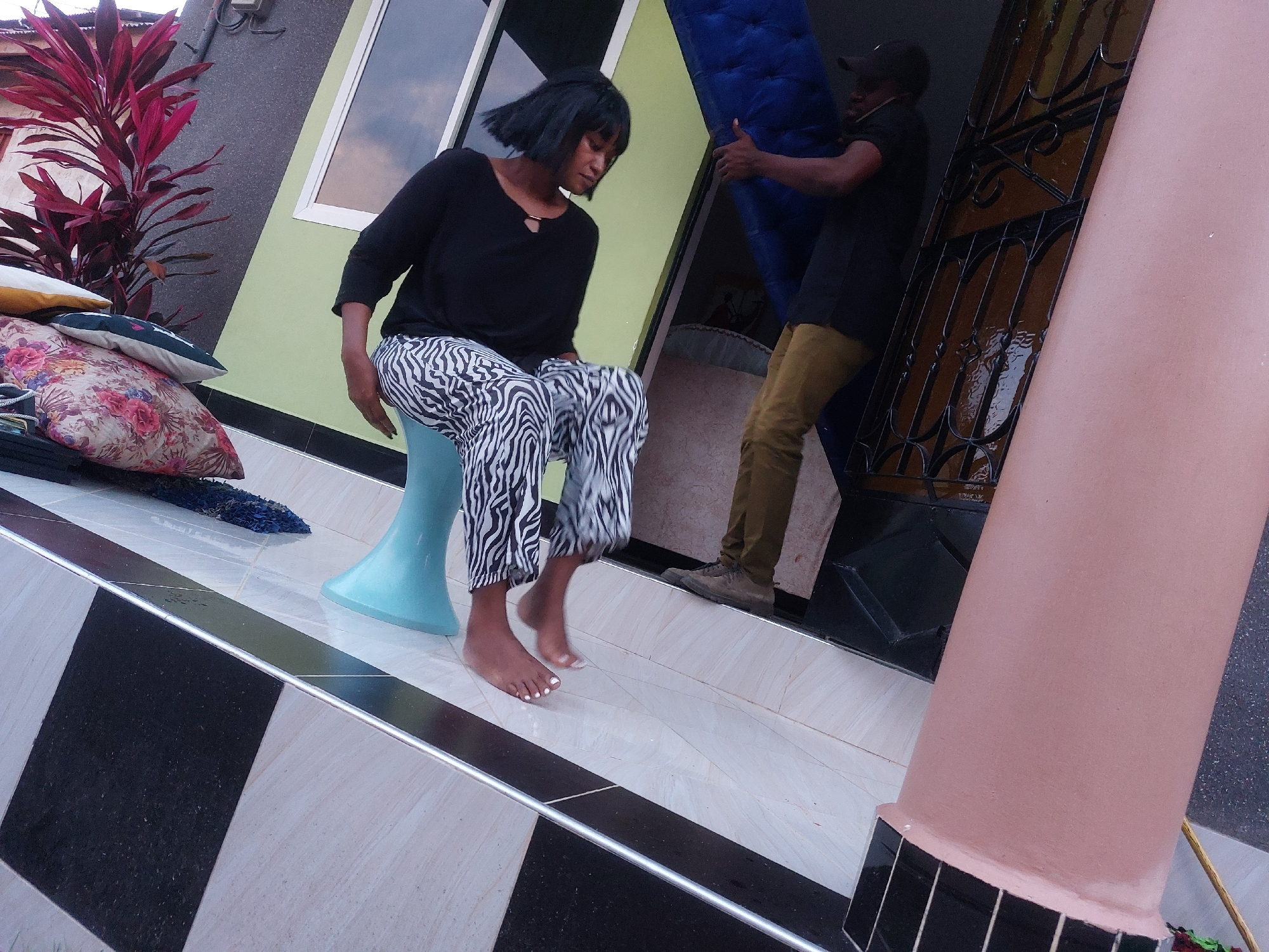 A woman is seated on a blue, cone-shaped stool on the tiled porch of a building, with a man behind her carrying a blue item. The woman has dark hair and is wearing a black long-sleeved shirt and zebra-print pants. She is barefoot, with white polish on her toenails. The man is wearing a black shirt, khaki pants, and a dark baseball cap. He is carrying what appears to be a rolled-up mattress or a similar large, soft object. Behind the woman, on the left, are several pillows stacked on top of each other, and a red leafy plant. A window is behind the woman, and an ornate, dark metal door is behind the man. The porch has black and white checkered tiles. The porch is next to a pink column. Based on the architectural style and vegetation, the location is suspected to be Dar es-Salaam, Tanzania.