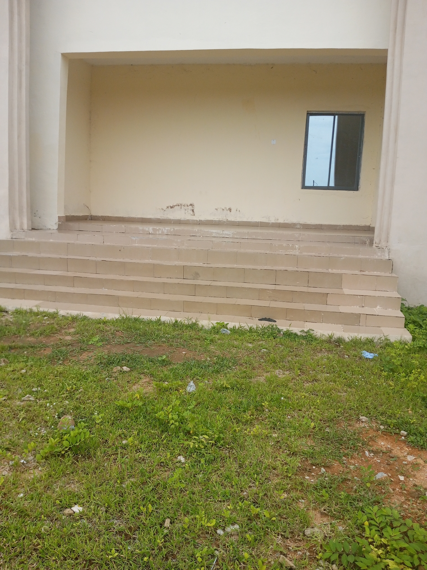 The image depicts the exterior of a building, possibly under construction or renovation, located in Jalingo, Nigeria. In the foreground, a patch of overgrown grass with some patches of bare earth suggests an unkempt outdoor area. Several small pieces of debris, including what appears to be plastic, are scattered on the ground.
Leading up to the building's entrance are a series of wide, shallow steps, approximately five or six in number, constructed from light-colored, possibly tiled, material. The steps appear clean and well-maintained.
The building itself has a light-colored, possibly cream or beige, facade. Above the steps, a recessed entrance area is visible, with a textured wall that shows some signs of wear, including discoloration and peeling paint. A single, small, rectangular window with a dark frame is situated on this wall. The windowpane reflects the sky. The building's exterior walls are partially obscured by large, fluted pilasters on either side of the entrance, also in a light, neutral color. The overall impression is of a simple, functional building in an outdoor setting.