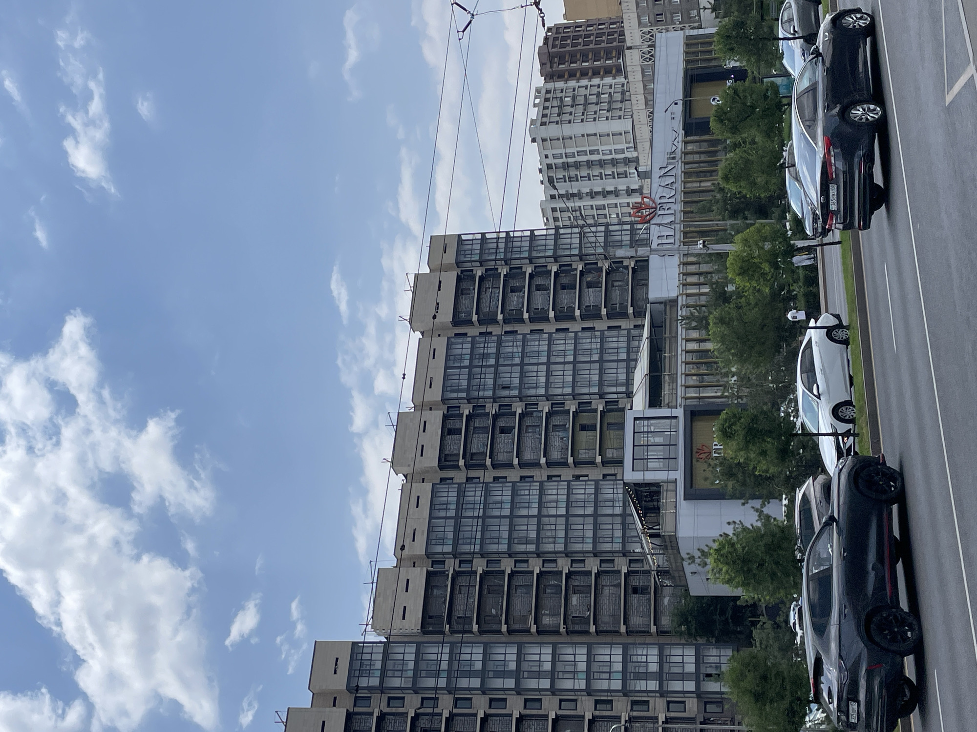 The image depicts a street scene in Almaty, Kazakhstan, with a large, modern residential building dominating the left side of the frame. The building features a facade of light-colored stone and numerous windows with dark frames, suggesting a contemporary architectural style. Partially visible in the background to the right are other buildings, including a white, multi-story structure and a brown, more traditional-looking building.
In the foreground, a paved road with lane markings is visible, with several cars in motion. A dark-colored SUV with red accents is in the left lane, while a dark-colored sedan is in the right lane. Other vehicles are also present, including a white sedan behind the SUV and a gray sedan further back.
Between the road and the buildings, there is a strip of greenery with trees. A commercial building with a prominent sign reading "HAFRAN" is situated in front of the residential building, partially obscured by trees. The sky above is partly cloudy, with patches of blue sky interspersed with white clouds. The overall scene appears to be a typical urban environment on a daytime.