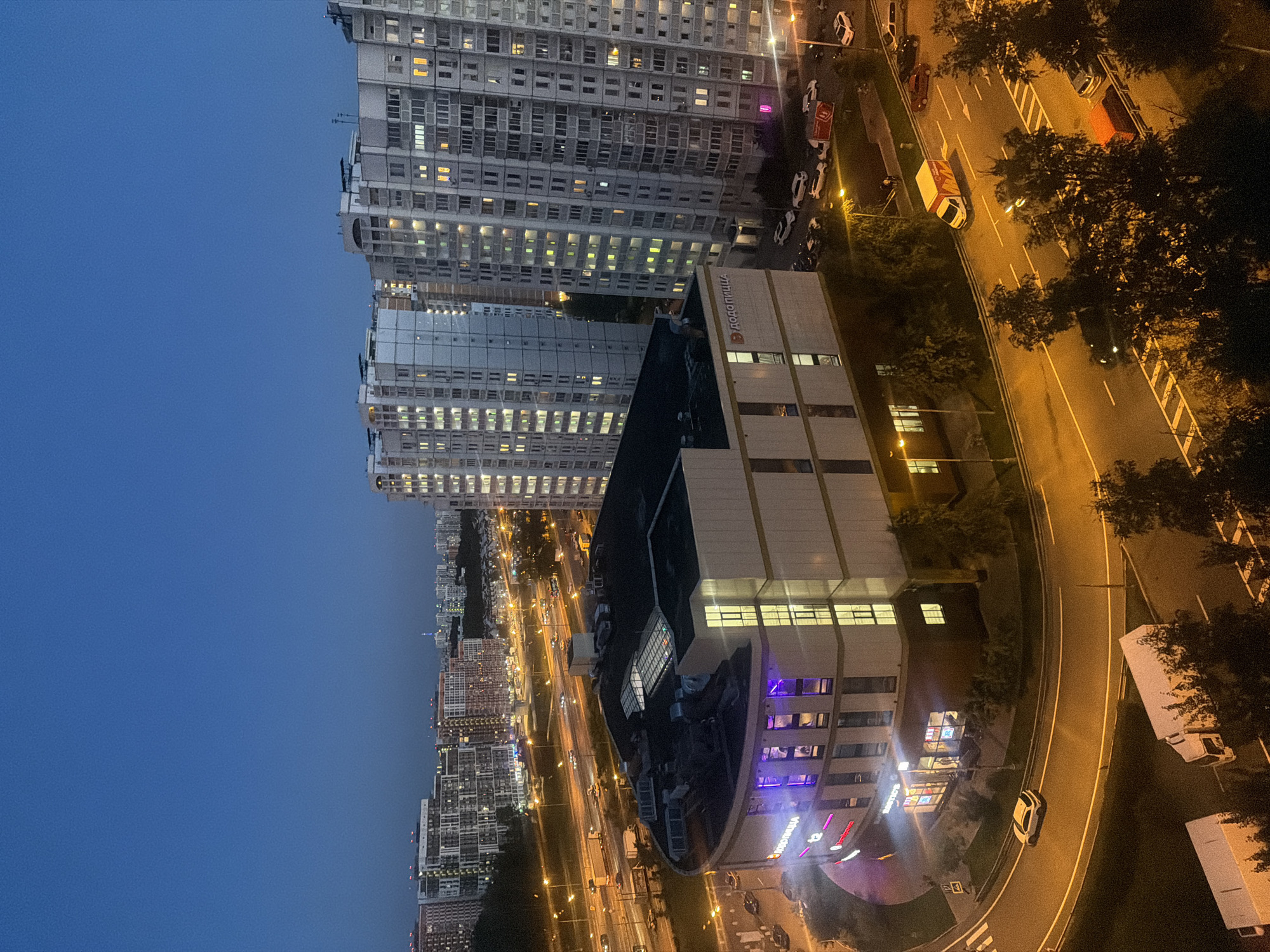 This is a high-angle, wide shot of a city at dusk. The sky is a deep twilight blue. In the foreground is a curved building with a white and beige facade. The building has many windows, some illuminated with purple and yellow light. A sign with Cyrillic text, appearing to be "Додо Пицца" (Dodo Pizza), is visible on the building. To the left of this building, a street curves around it. A white car is driving on this curved street, and a crosswalk is visible.
Behind the curved building, a multi-lane highway stretches into the distance, illuminated by streetlights and the lights from vehicles. Various cars and trucks are visible on the highway. Further in the background, numerous tall apartment buildings are densely packed together. These buildings are illuminated with many yellow and white lights, indicating occupancy. The overall scene depicts a residential and commercial area of a city at night, with active traffic on the highway. The location is identified as Дюна Сск, Russia.