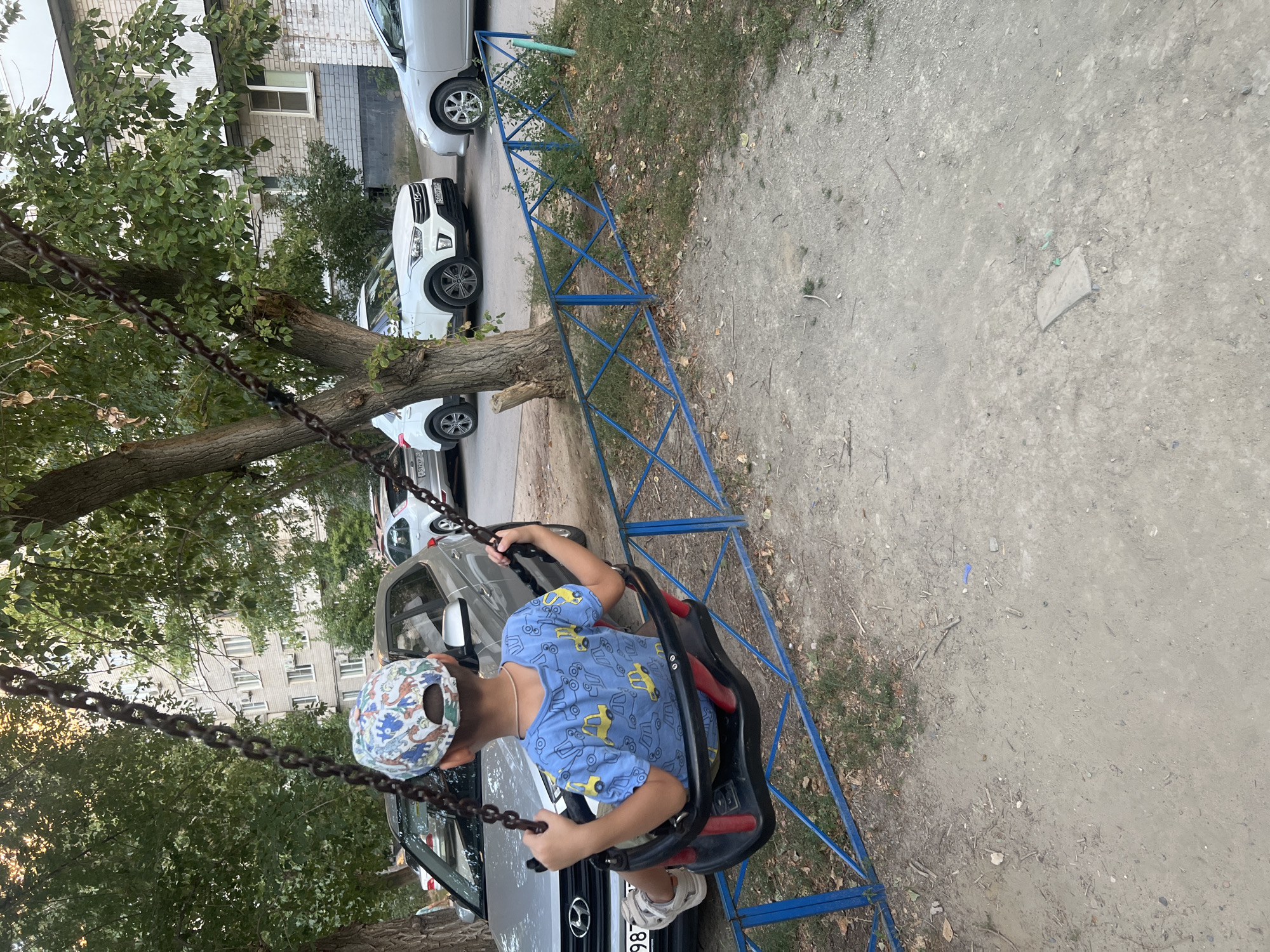 A young boy, facing away from the camera, is sitting on a swing in a playground. He is wearing a blue t-shirt with a yellow car print and a white and blue patterned baseball cap. The swing set is made of metal chains and has a black plastic seat with red accents. The playground is located in front of a multi-story apartment building in Volgograd, Russia. Several cars are parked on the street behind the swing set, including a silver Hyundai and a white SUV. A blue metal fence separates the playground from the parking area. The ground is a mixture of dirt and gravel. The time of day appears to be late afternoon, with the sun casting long shadows.