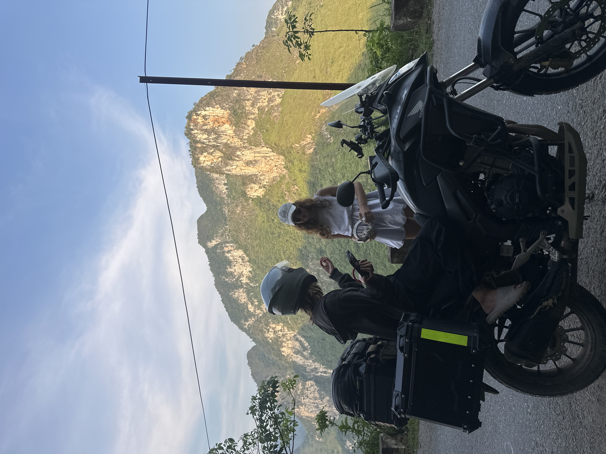 The scene is an outdoor setting in Đường Thượng Commune, Vietnam, with a backdrop of a large, weathered mountain covered in vegetation under a partly cloudy blue sky. A black motorcycle is parked on the right side of the frame, partially obscuring the view. Two people are present. One person, wearing a helmet and dark clothing, is seated on the motorcycle and appears to be looking at a mobile phone. Another person, dressed in a light-colored dress and cap, stands beside the motorcycle. The individual in the dress is holding a cylindrical object. A black storage container is attached to the back of the motorcycle. A utility pole stands in the background on the left side. Vegetation is also visible near the bottom left corner.