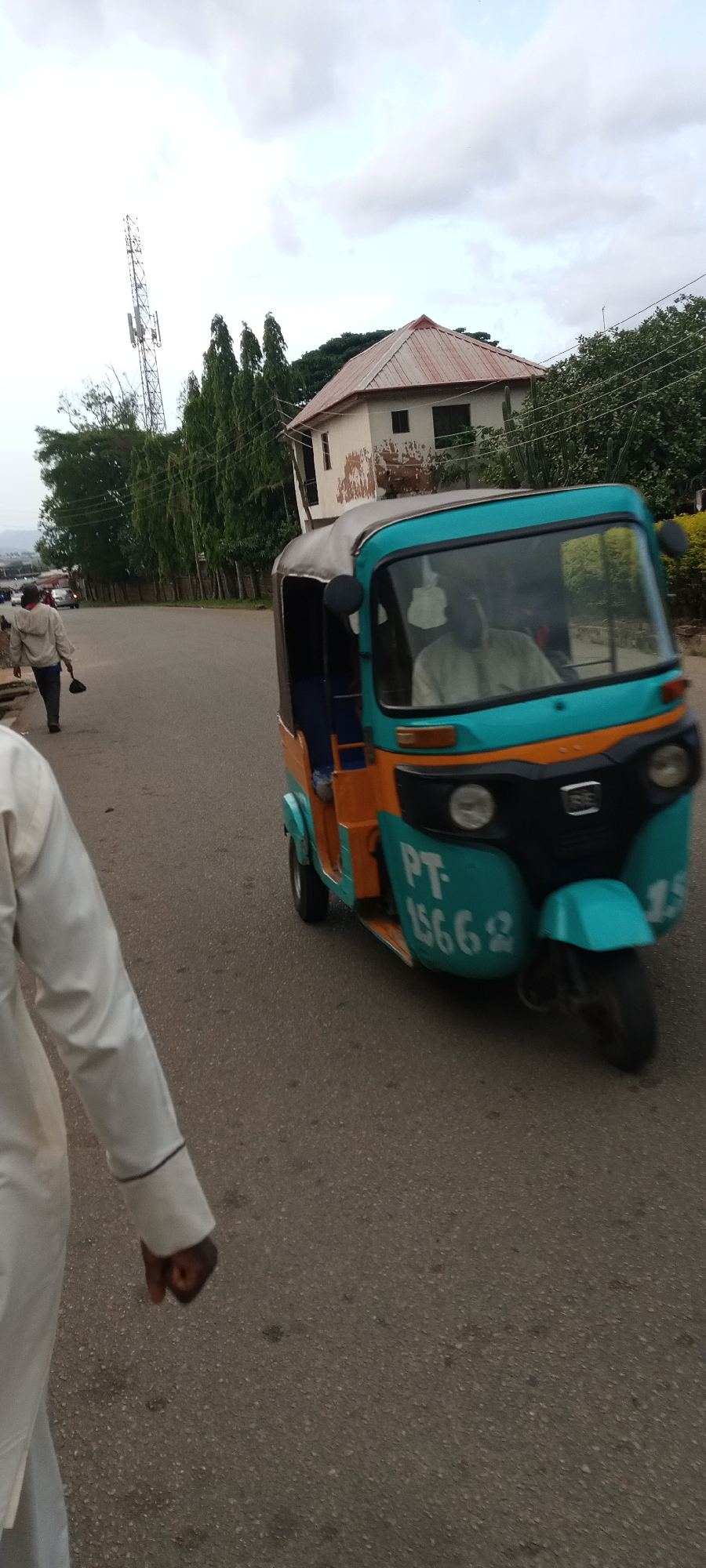 The image shows a street scene in Jos, Nigeria. A turquoise and orange auto-rickshaw, commonly known as a "keke," is moving along the road. The keke has the registration number "PT-15662" on its front. An individual is driving the keke. A man in a white traditional gown is walking along the side of the road. Another man can be seen walking further down the road, holding a broom-like object. A building with a rusty red roof is visible in the background, along with some trees and a communications tower. The road is paved and appears to be moderately busy.