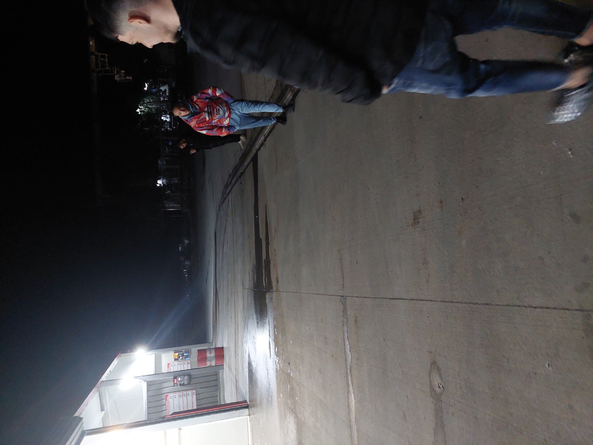 The image shows an outdoor scene at a gas station, likely in Asenovgrad, Bulgaria. The foreground is dominated by a concrete surface, possibly a driveway or parking area, showing signs of recent moisture or cleaning. A person wearing blue jeans and a dark jacket is partially visible in the top center of the image, moving through the frame. Another person is seen wearing a patterned red jacket and blue jeans. In the background, a gas station structure is illuminated, with price displays and signage visible. A dark area suggests a night setting. Other individuals are partially visible in the background.