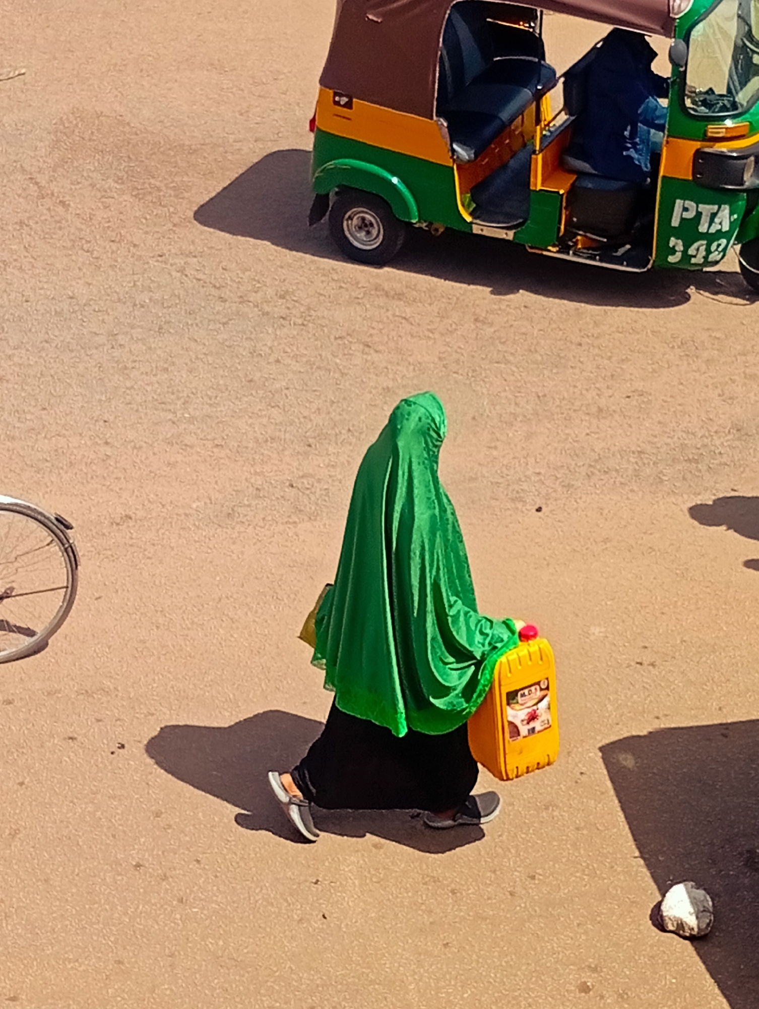 In the city of Bukuru, Nigeria, a person walks across a dusty street, partially obscured by a bright green hijab. They are carrying a yellow plastic container, likely filled with liquid, in their right hand. The container has a red cap and a label with some text and an image. The person is wearing dark pants and dark shoes with white accents. Behind them, a colorful three-wheeled auto-rickshaw is parked on the street. The auto-rickshaw is predominantly green and yellow with a brown canopy and has license plates that read "PTA 342". A partial view of a bicycle wheel is visible on the left side of the image. The ground is paved with a light brown, textured material, and shadows are cast by the objects on the street, indicating a sunny day.